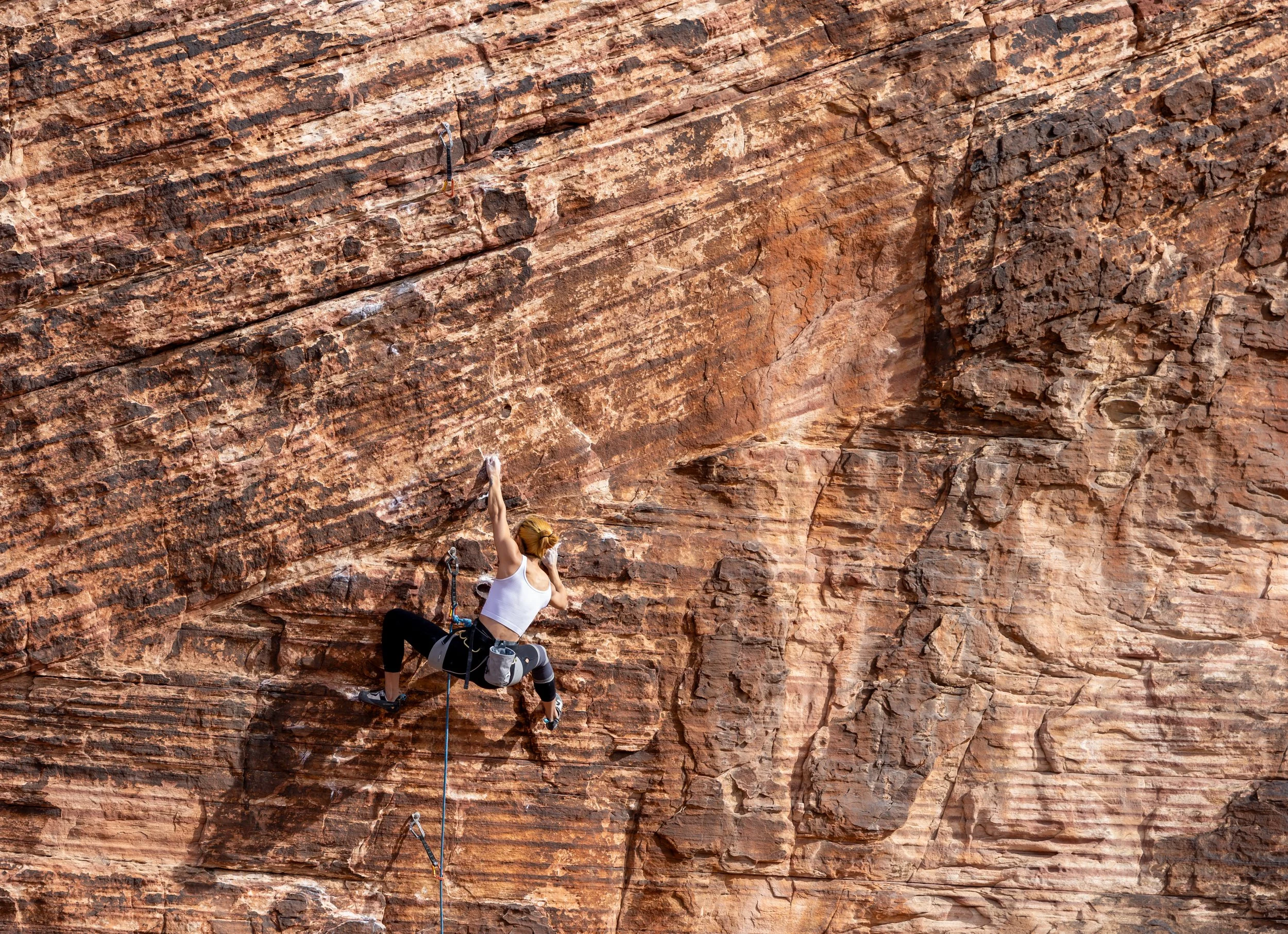 A woman rock climbing on a large red and brown sandstone cliff, wearing climbing gear and reaching up with her right hand.
