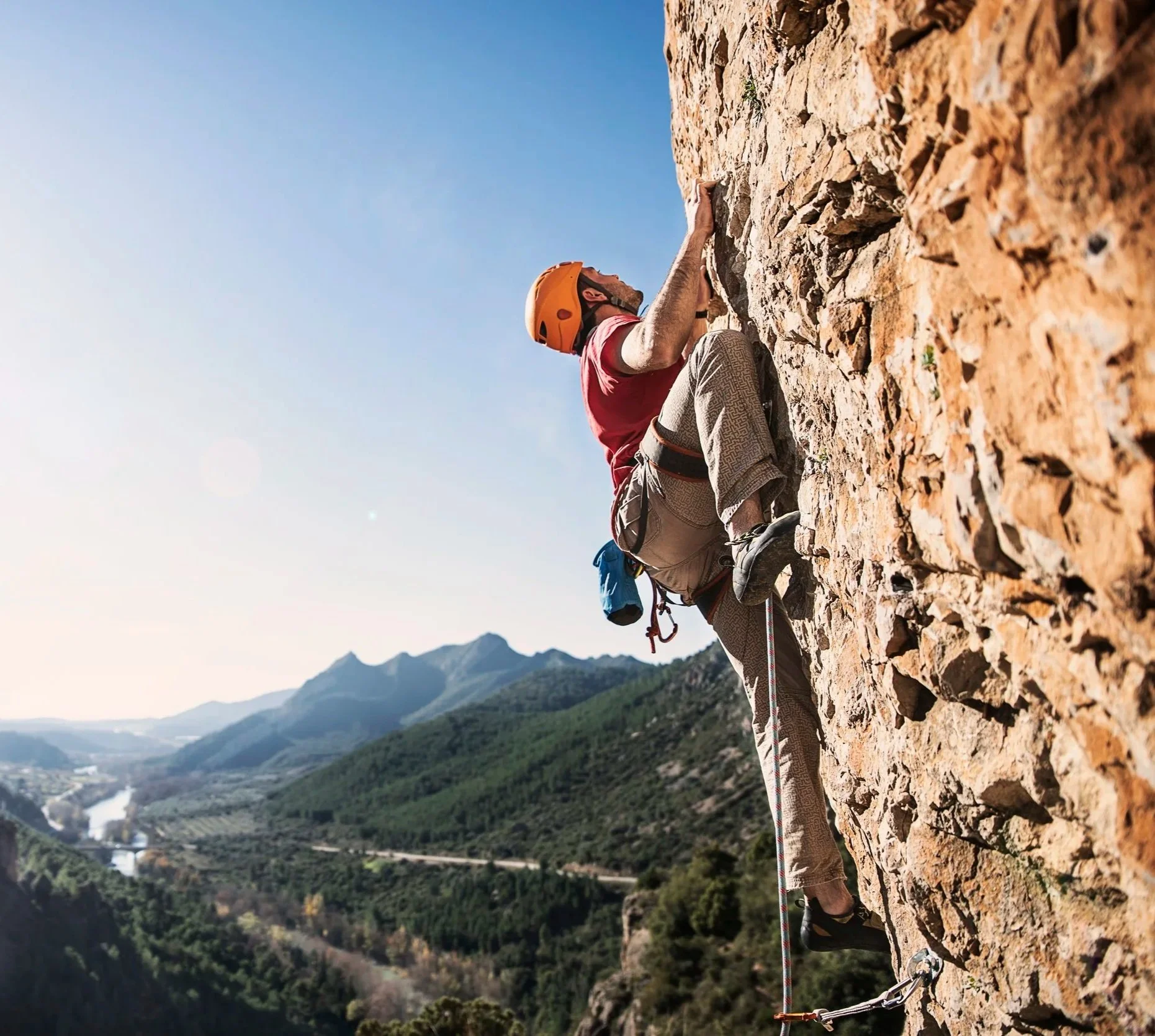 A male rock climber wearing a yellow helmet and red shirt scaling a steep, rocky cliffside with a scenic valley and mountains in the background.