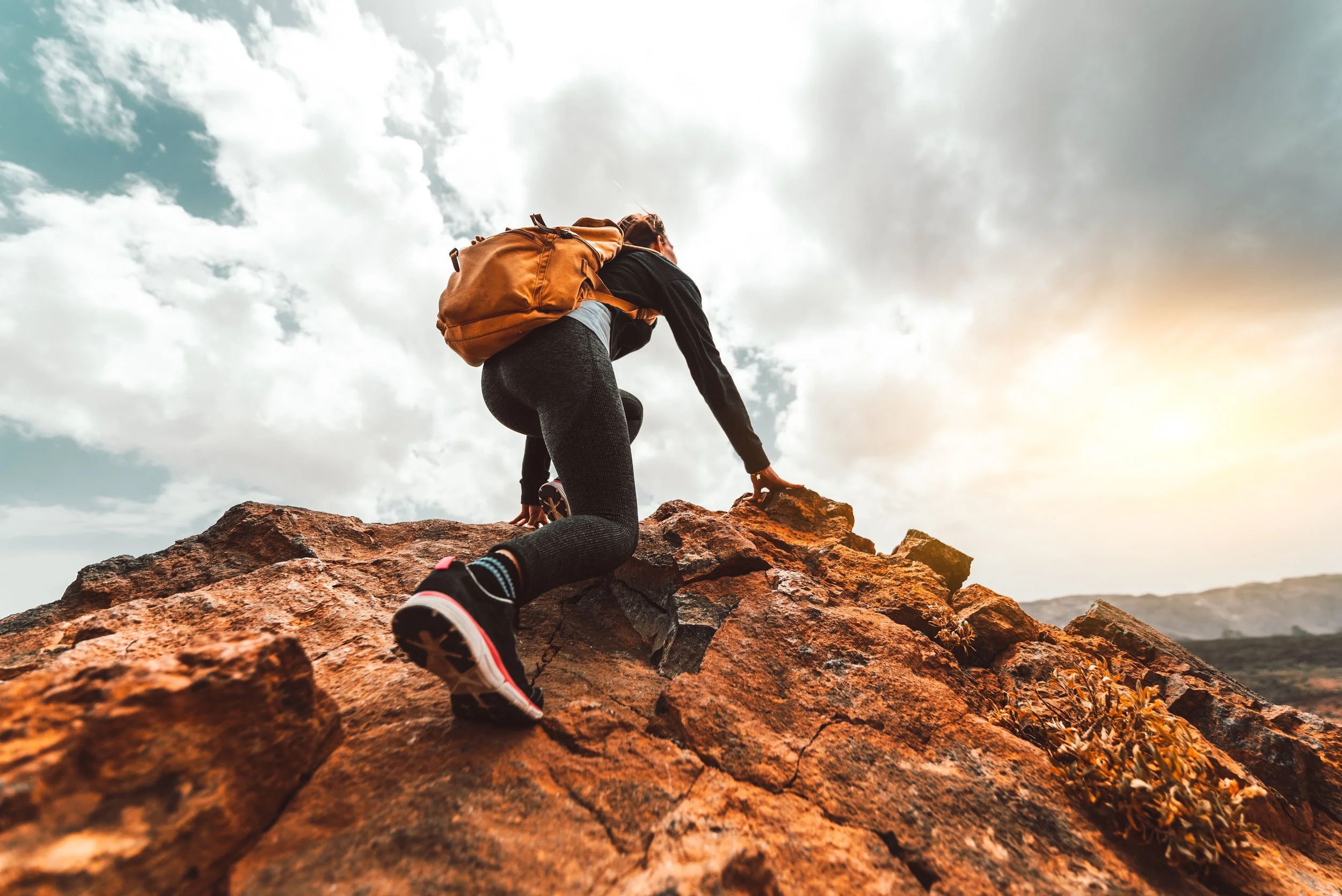 A person climbing a rocky terrain outdoors with a backpack, with a cloudy sky and sunlight in the background.