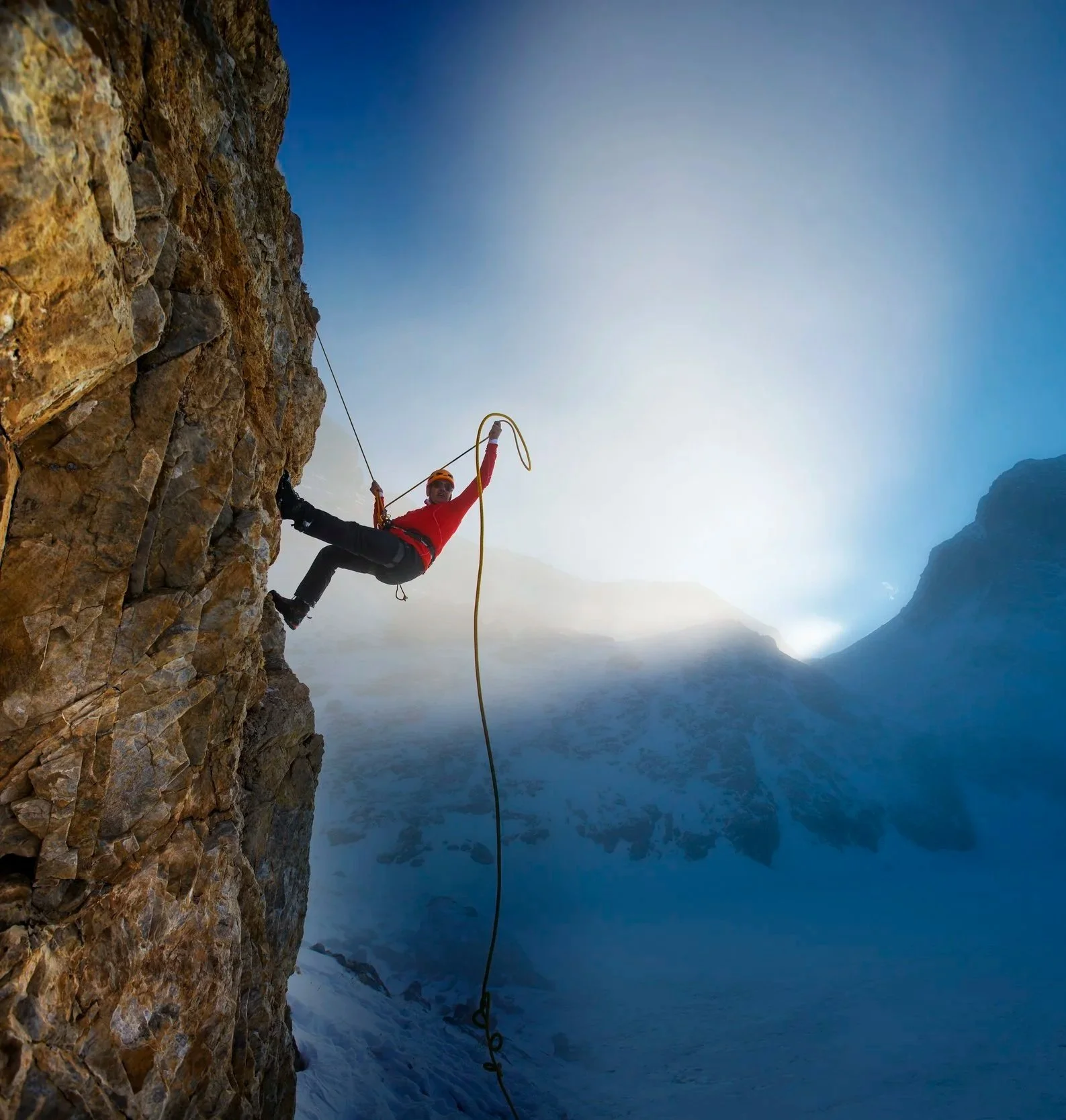 A person dressed in climbing gear, including an orange helmet and red jacket, is rappelling down a steep rocky cliff in a mountain landscape covered with snow, with mist and mountains in the background.