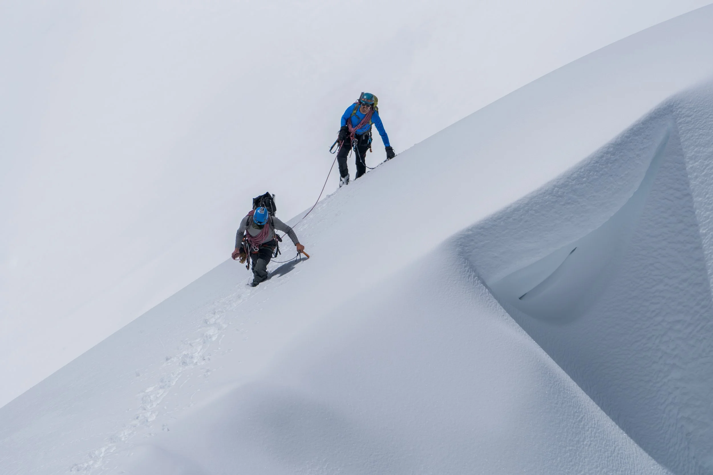 Two climbers ascending a snowy mountain slope with trekking poles, wearing helmets and winter gear.