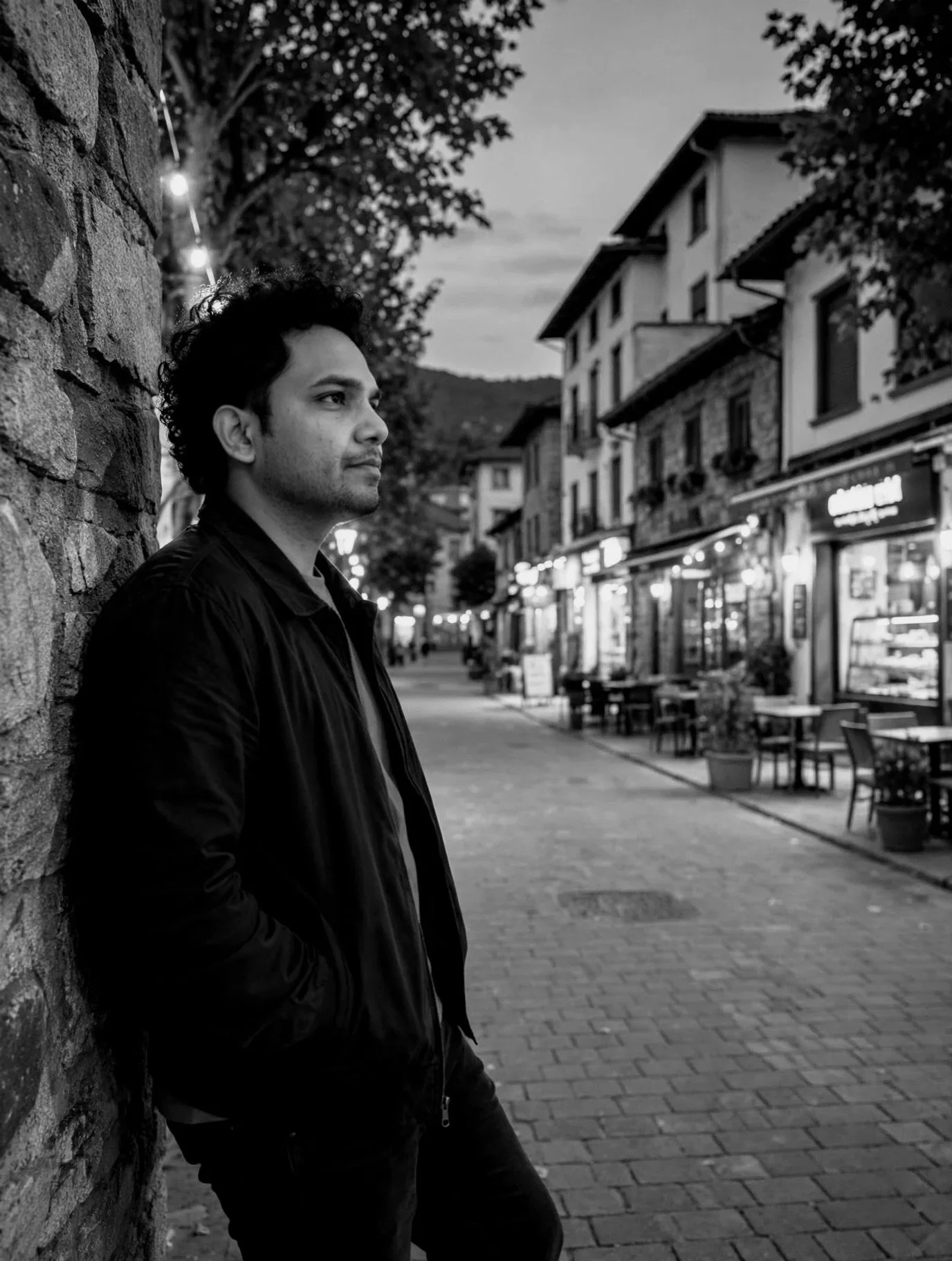 Vejeps Ephi Kingsly with curly hair and a light beard standing on a city street at dusk, leaning against a brick wall and gazing into the distance. The street is lined with shops and outdoor tables.