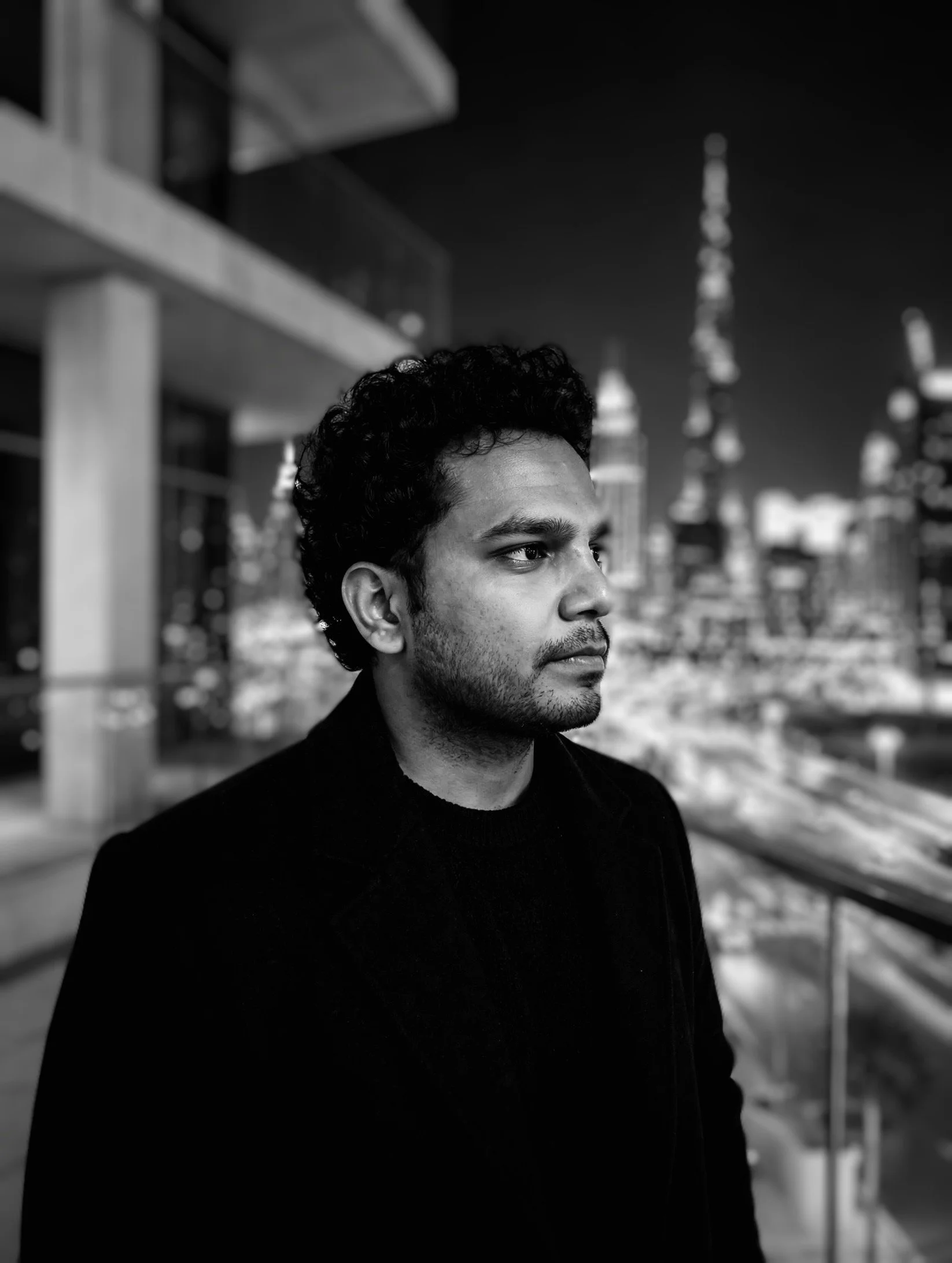 A black and white photo of a man with curly hair and a beard, wearing a dark coat, standing outdoors at night with city lights and buildings in the background.
