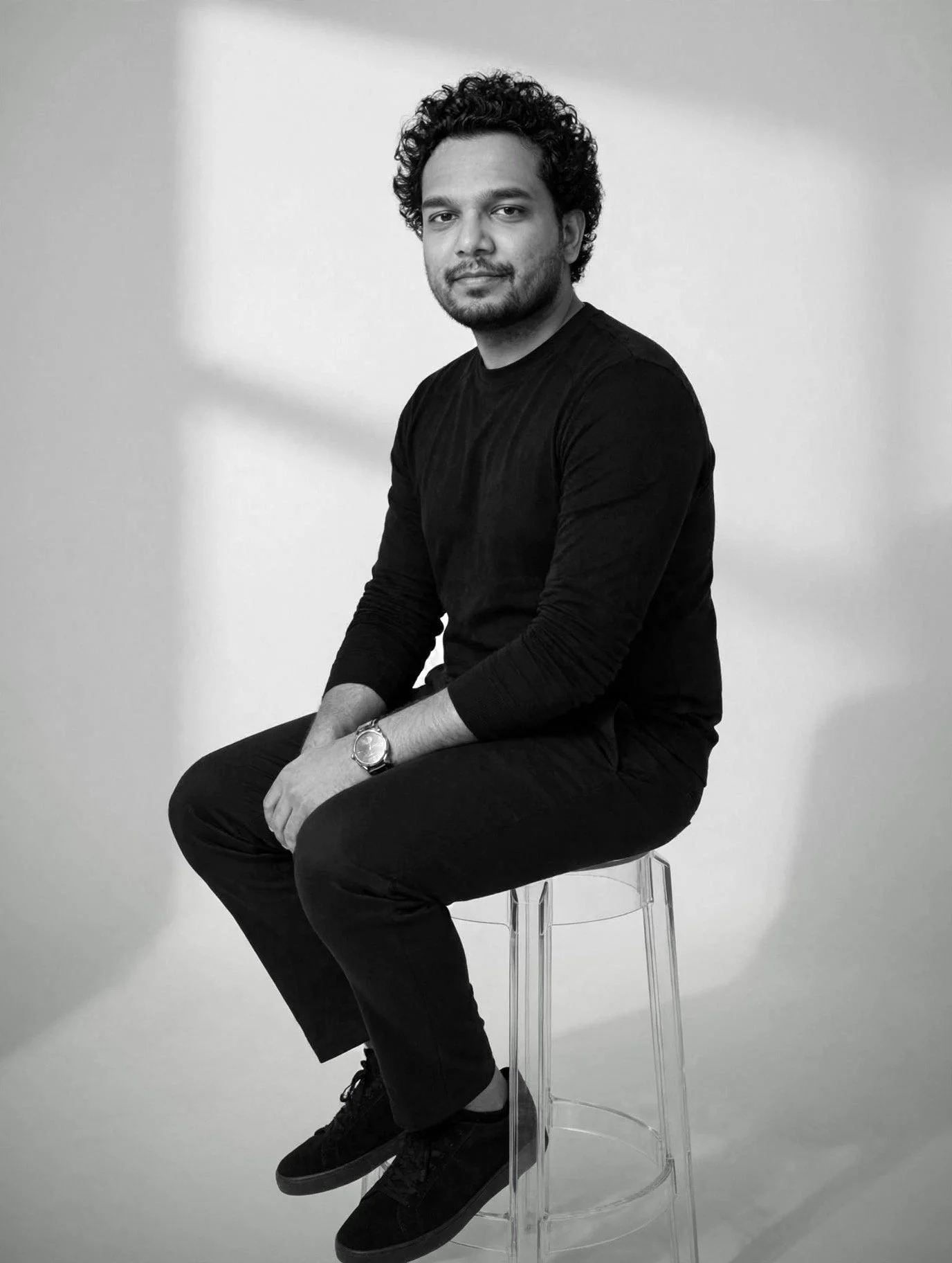 A man with curly hair and a beard wearing a black long-sleeve shirt and black pants seated on a transparent stool in front of a plain white background.