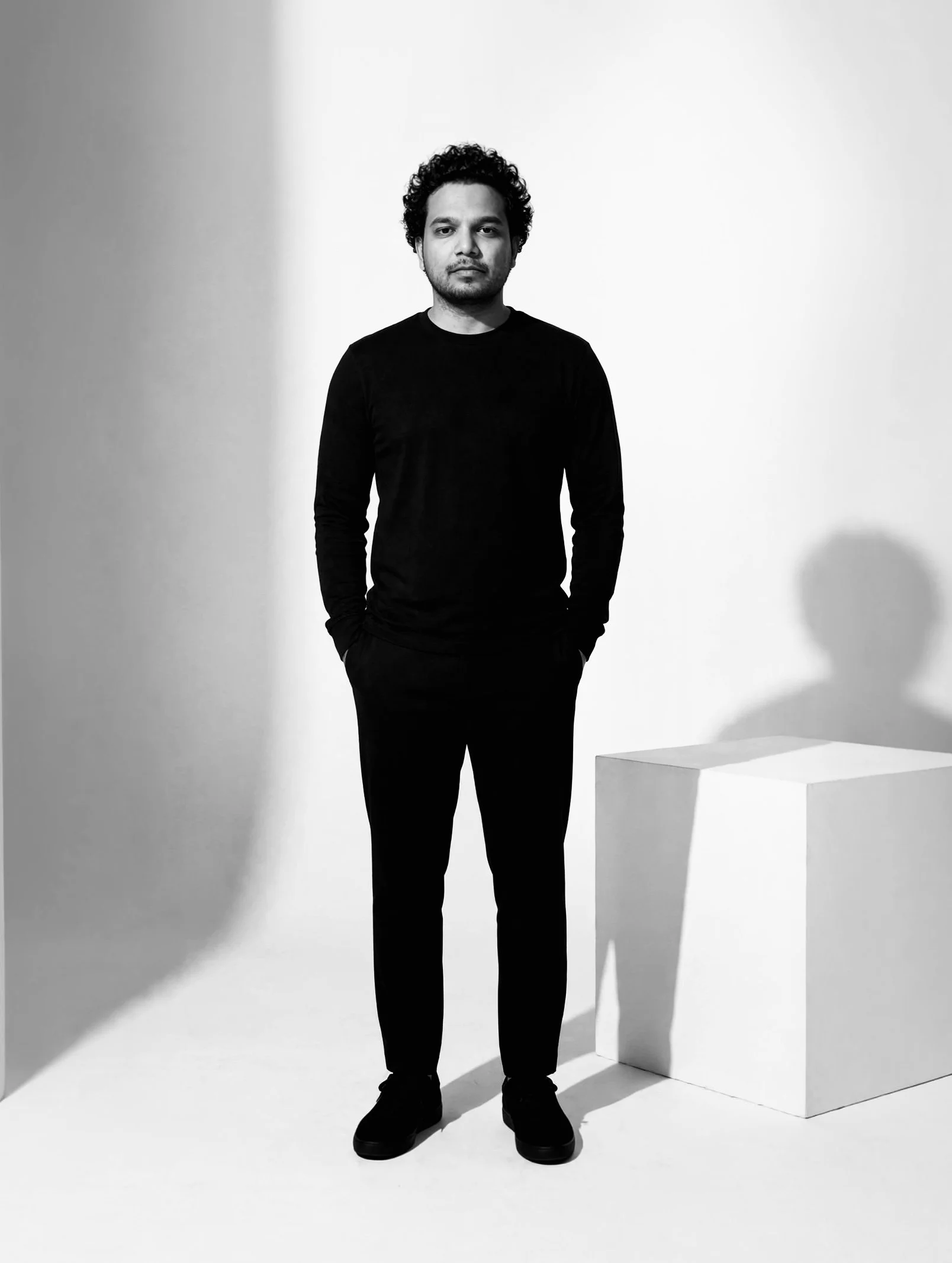 Black-and-white photograph of Vejeps Ephi Kingsly  standing in a minimalist studio with high contrast lighting, wearing a black long-sleeve shirt and black shoes, with a white geometric sculpture beside him.