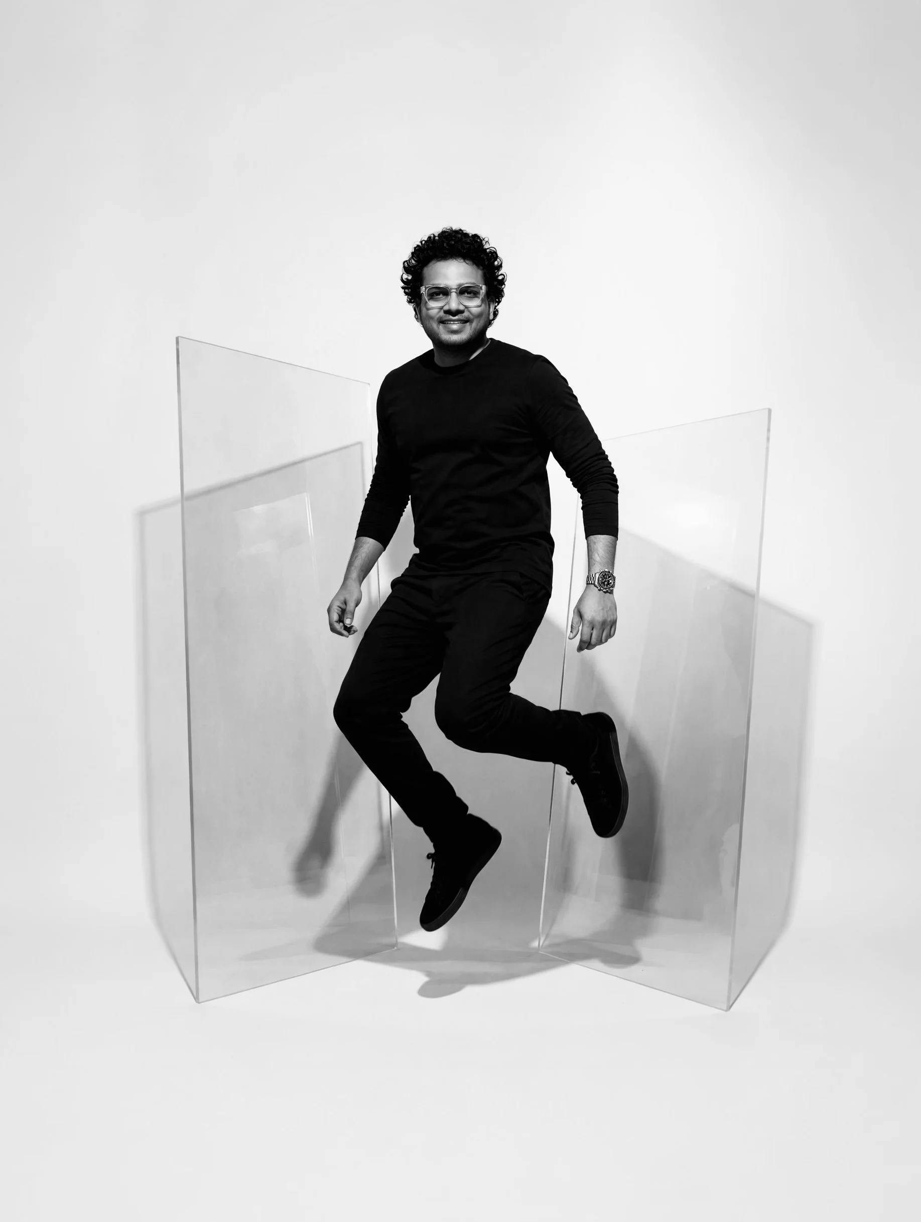A man with curly hair, glasses, and a watch is sitting on a clear acrylic chair in front of a white background.