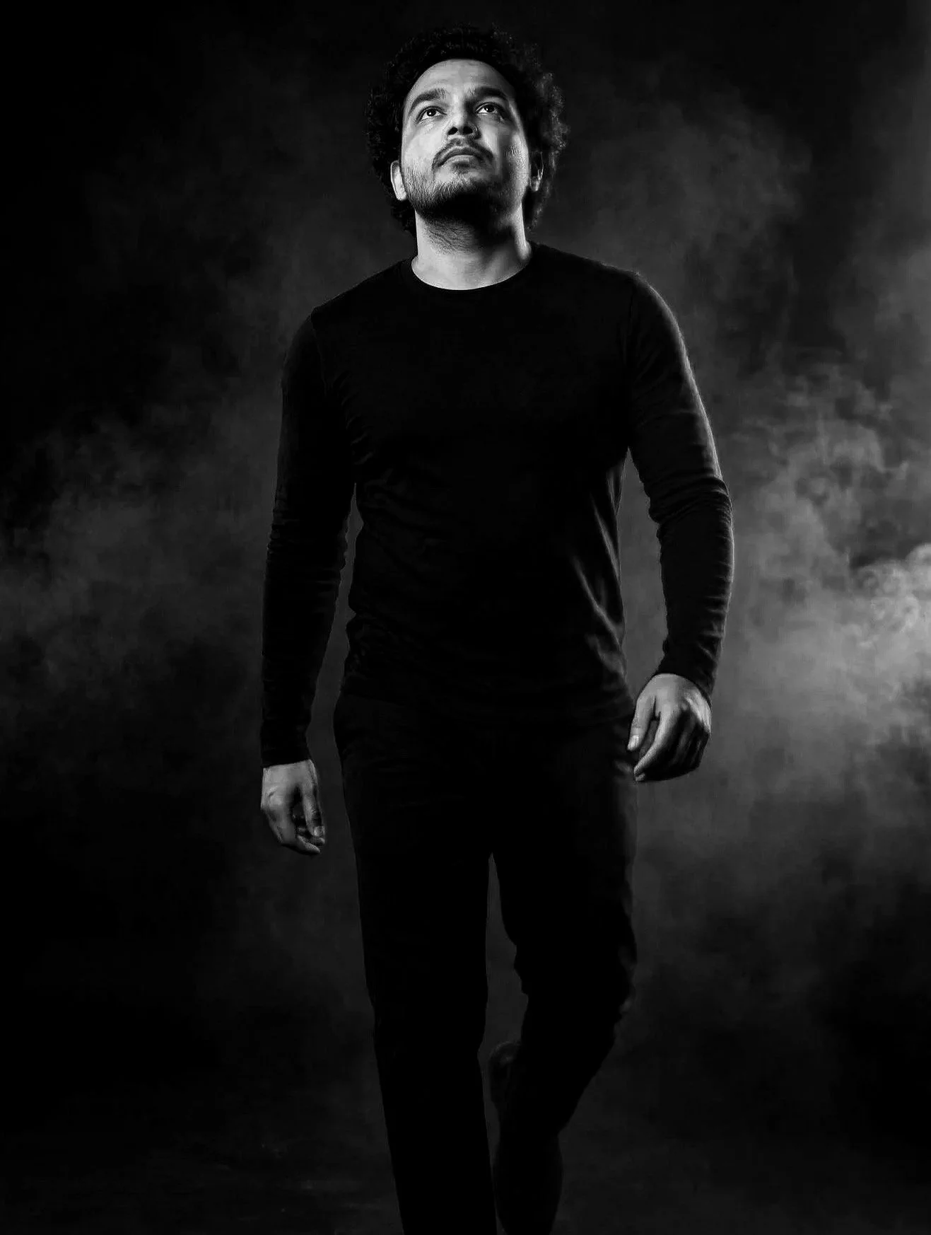 A black and white portrait of a man with curly hair, looking upwards, dressed in a long-sleeved shirt, walking against a dark, misty background.