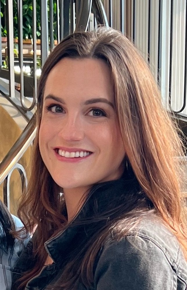 A young woman with long wavy brown hair and a big smile, wearing a black leather jacket, sitting outdoors near a metal railing.