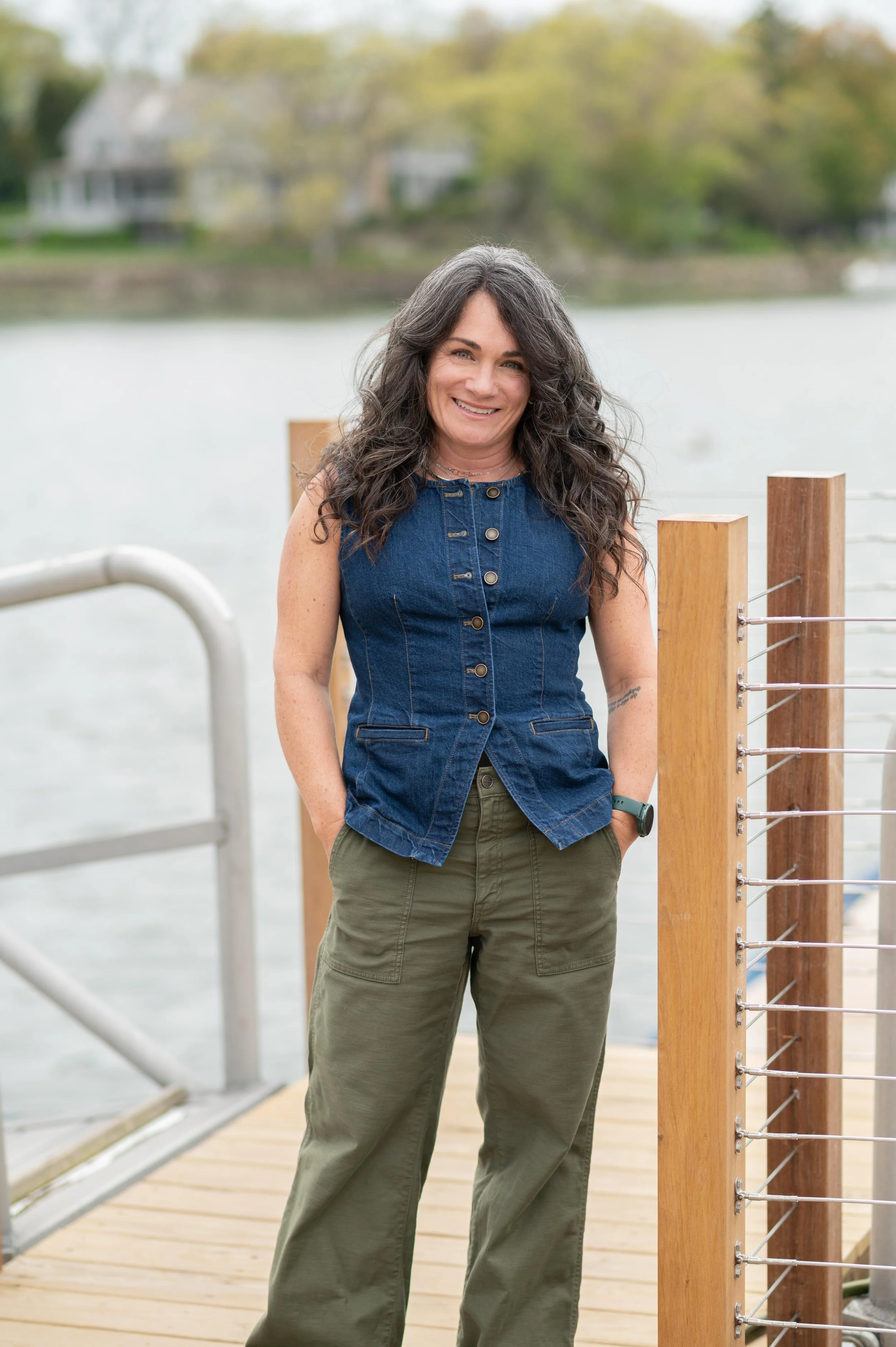 Woman with long curly gray hair smiling, wearing a sleeveless denim button-up top and olive-green cargo pants, standing on a wooden dock near water with houses and trees in the background.