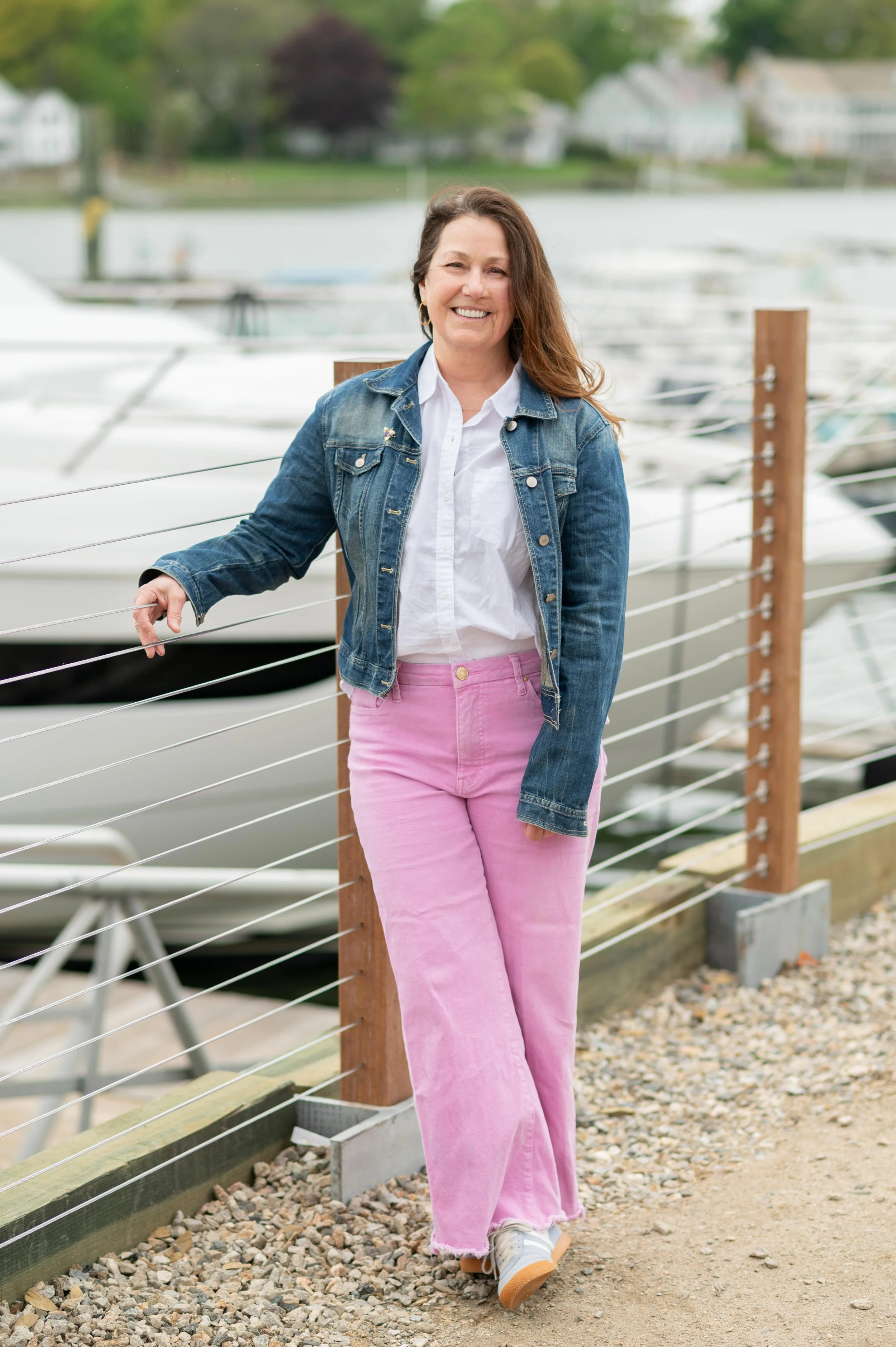 A smiling woman with long brown hair, dressed in a denim jacket, white shirt, and pink pants, walking along a marina with boats and greenery in the background.