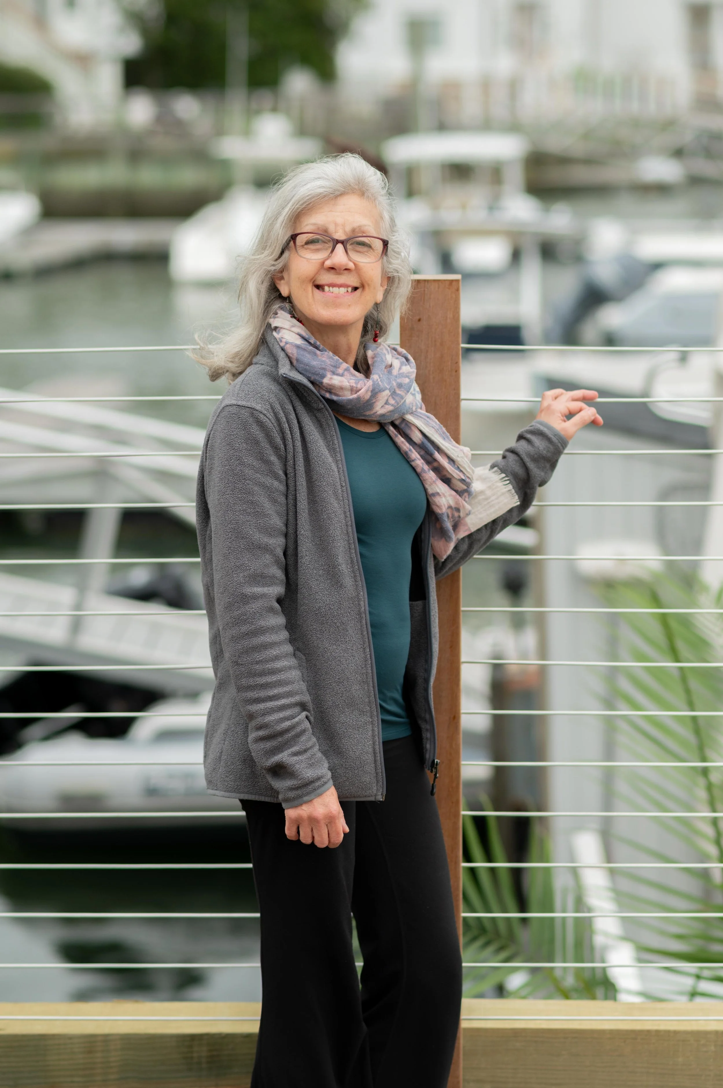An older woman with gray hair and glasses smiling while standing outdoors near a marina with boats and water in the background.
