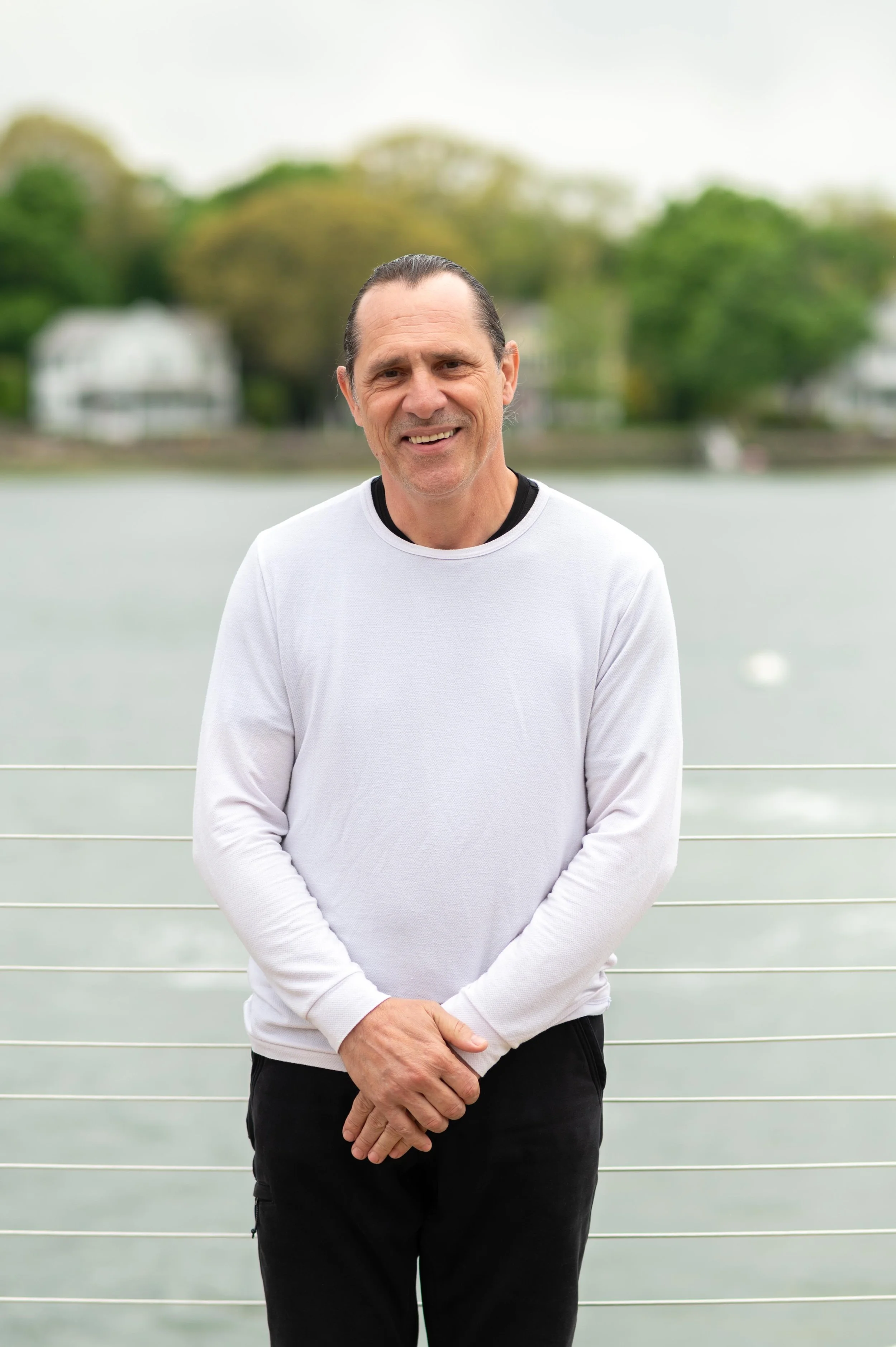 A man with slicked back hair wearing a white long-sleeve shirt and black pants, standing outdoors in front of a body of water with green trees and houses in the background.