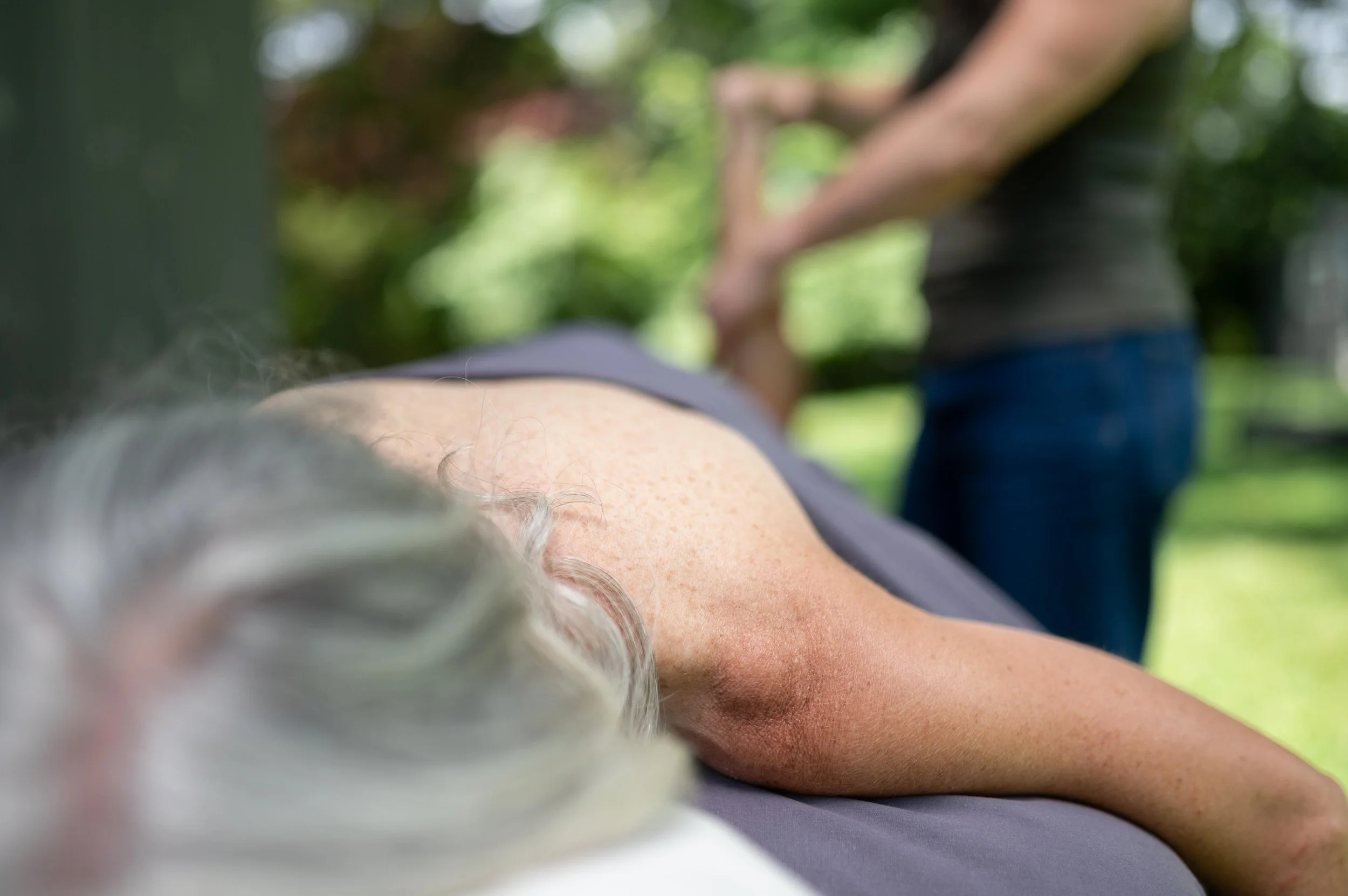 A close-up photo of a person with gray hair lying on a surface outdoors, with another person and blurred greenery in the background.