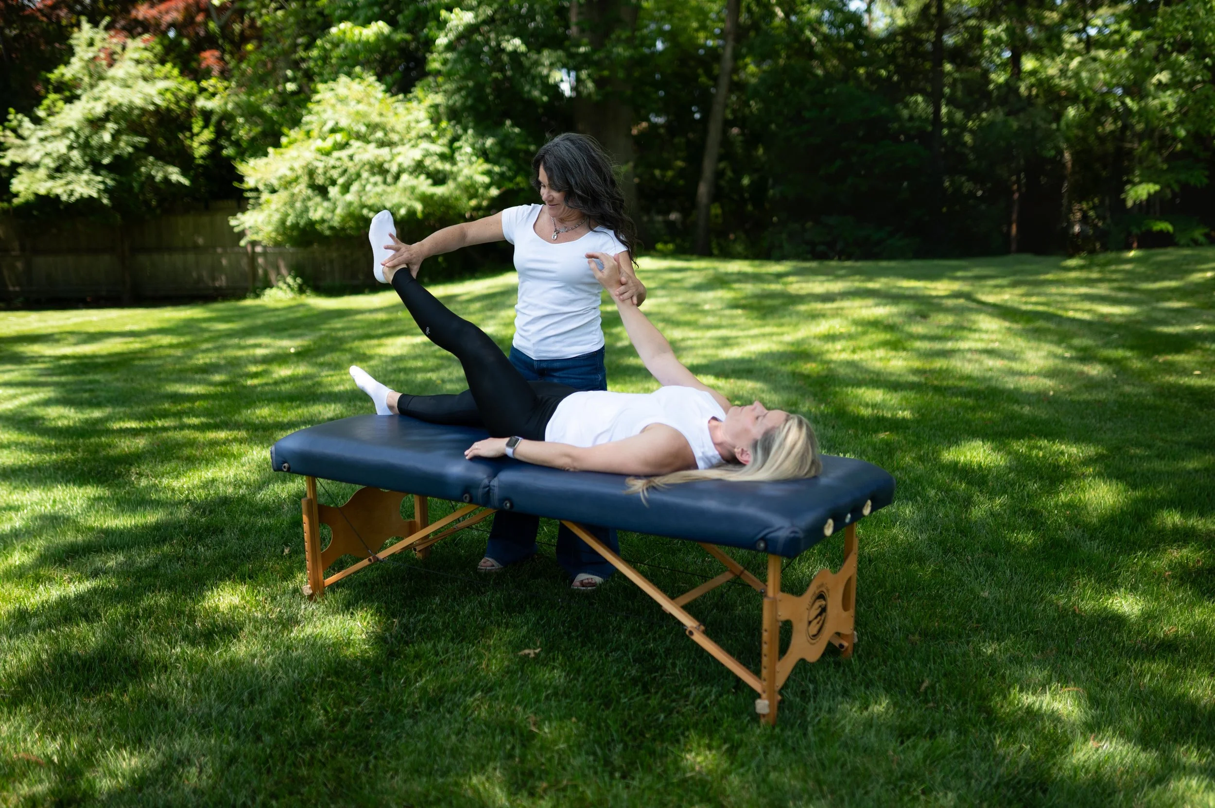 A woman lying on a portable massage table outdoors in a grassy backyard with trees, receiving a leg massage from another woman standing beside her.