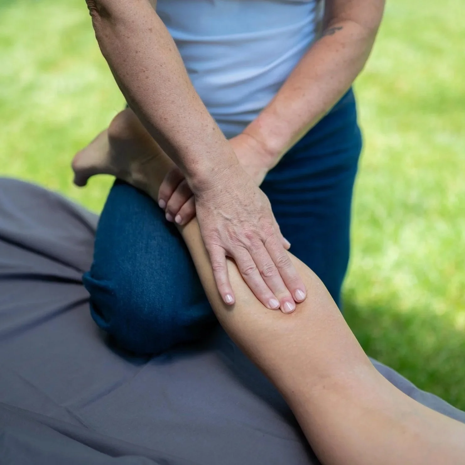 A person receiving a massage or physical therapy on their leg outdoors, with a massage therapist kneeling and applying pressure with their hands.