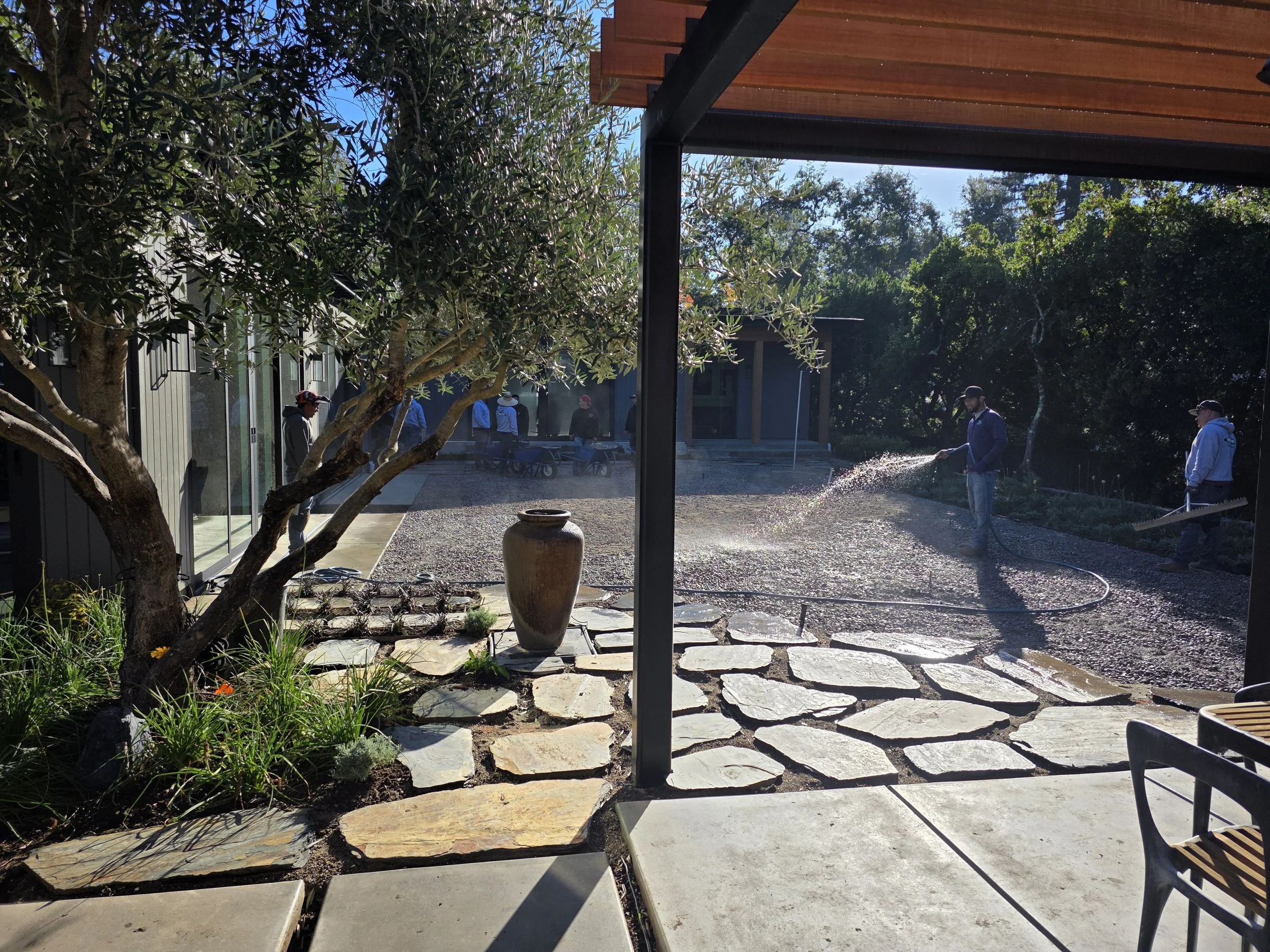 People working outdoors in a garden or yard, some wearing hats, with trees and buildings in the background, and water spraying from a hose.