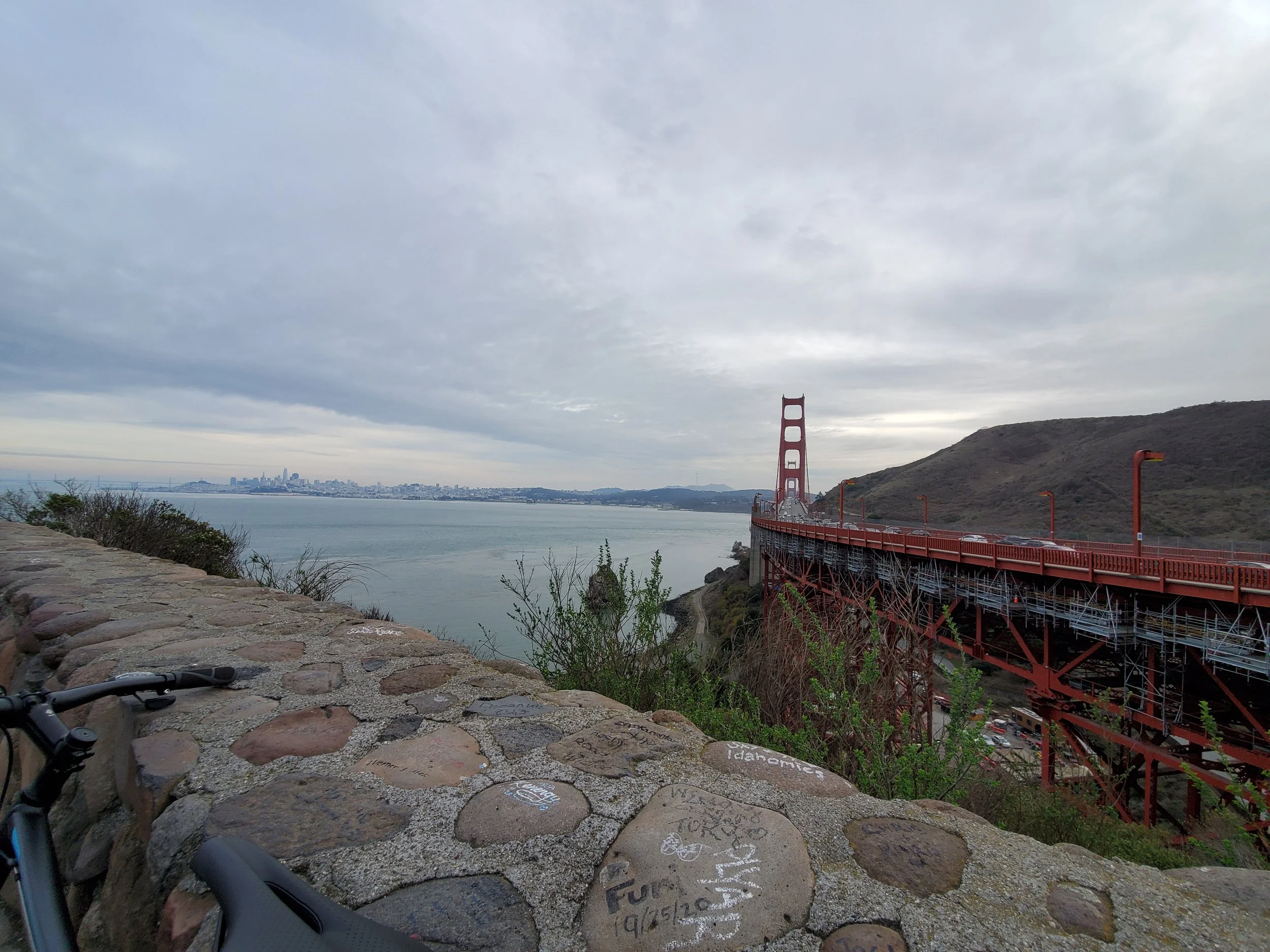 View of the Golden Gate Bridge in San Francisco under cloudy skies, with the city skyline in the background and a bicycle handlebar in the foreground.