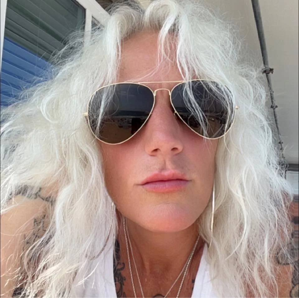Close-up of a woman with curly white hair wearing aviator sunglasses and a white tank top, outdoors with a building and window in the background.