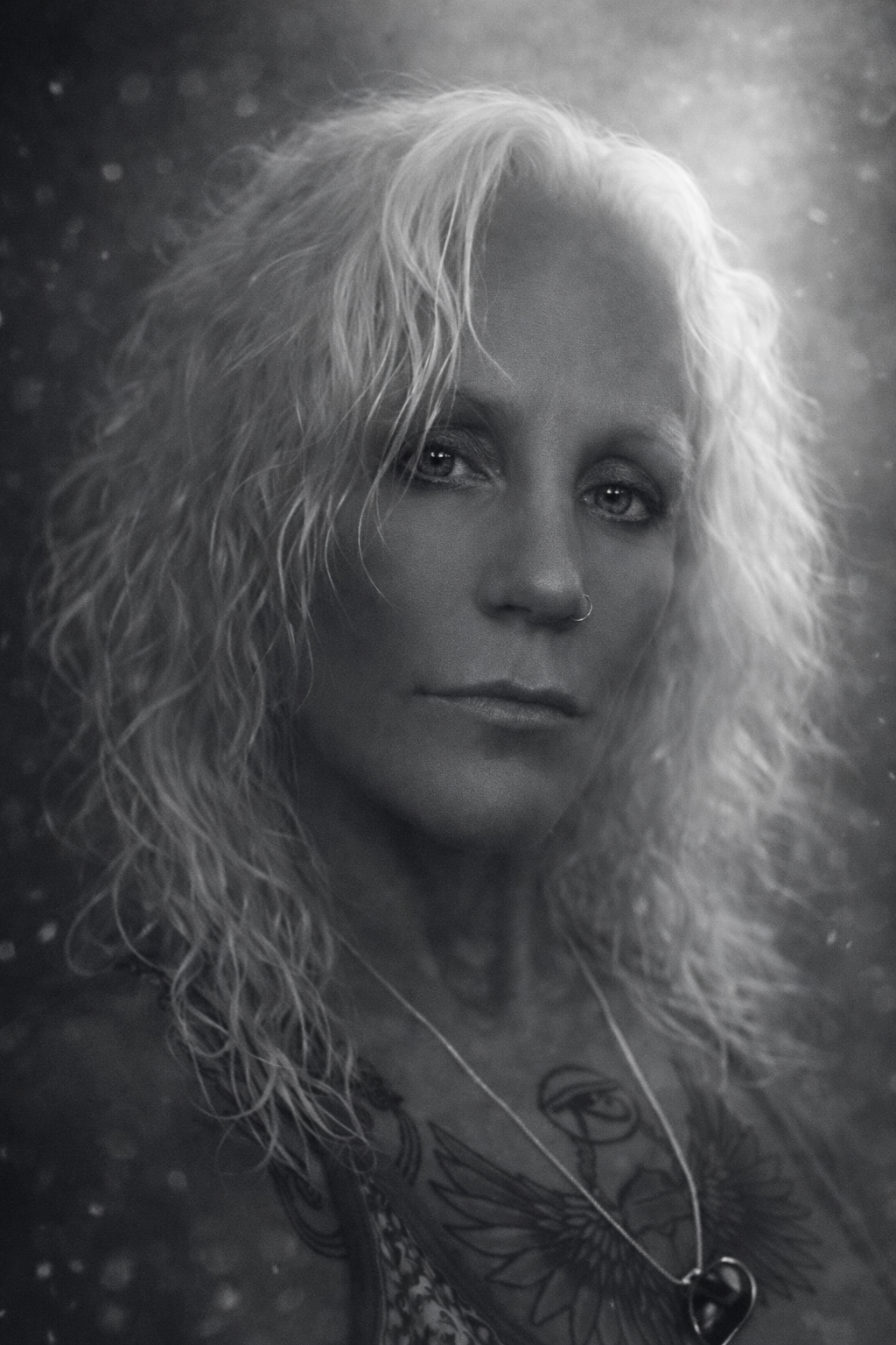 Black and white portrait of a woman with curly blonde hair, wearing a necklace with a heart-shaped pendant, and a nose ring, looking directly at the camera.