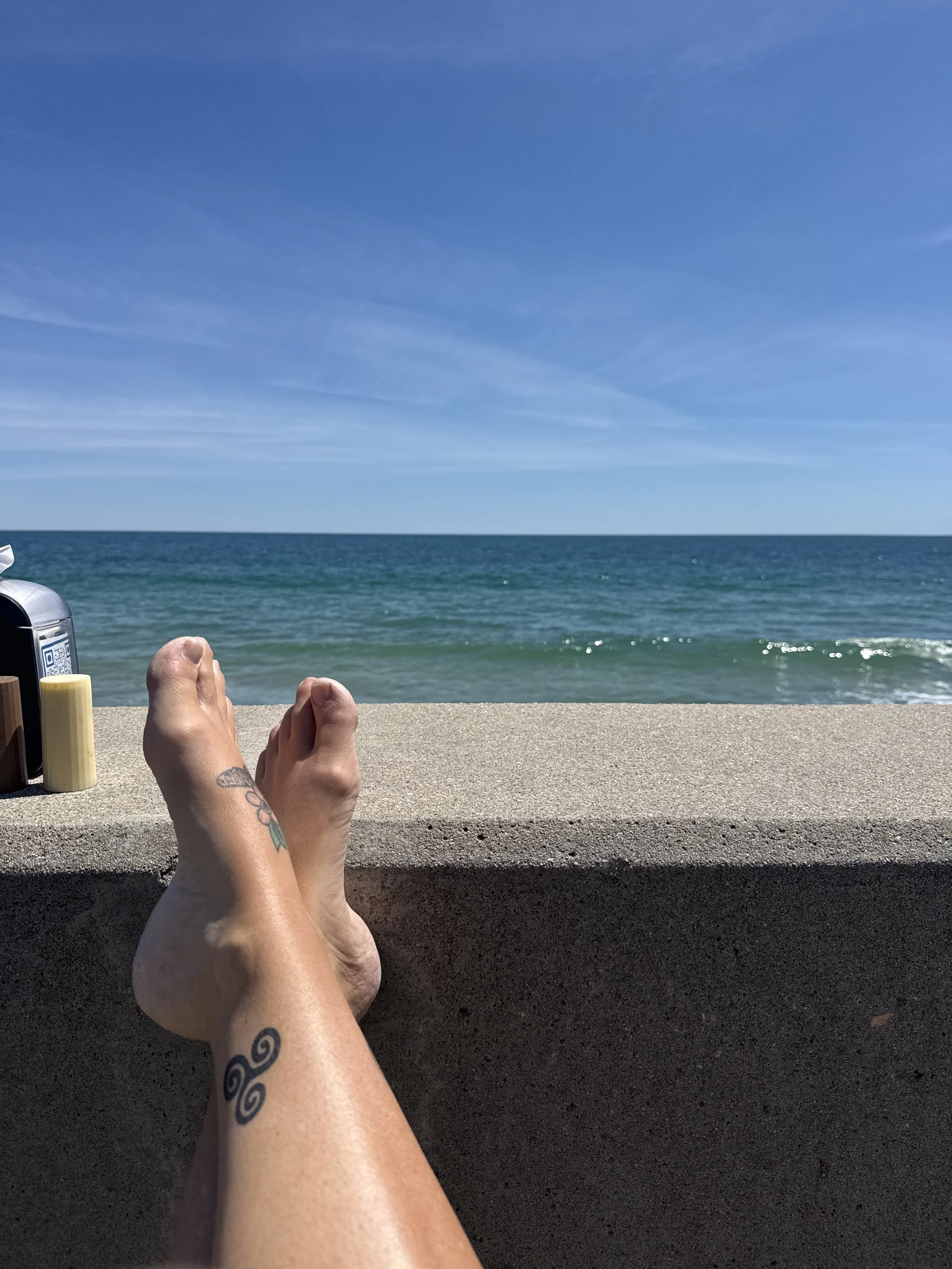 Person relaxing on a beachside concrete structure with their feet outstretched, overlooking the ocean and a clear blue sky.