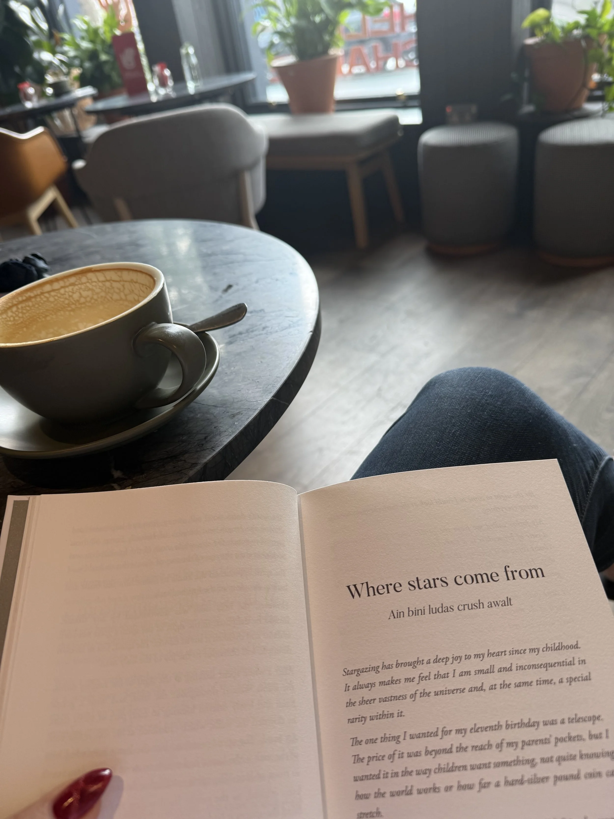 A person sitting in a cafe, with a coffee cup on a marble table, an open book with visible text, and several chairs and potted plants near the window.
