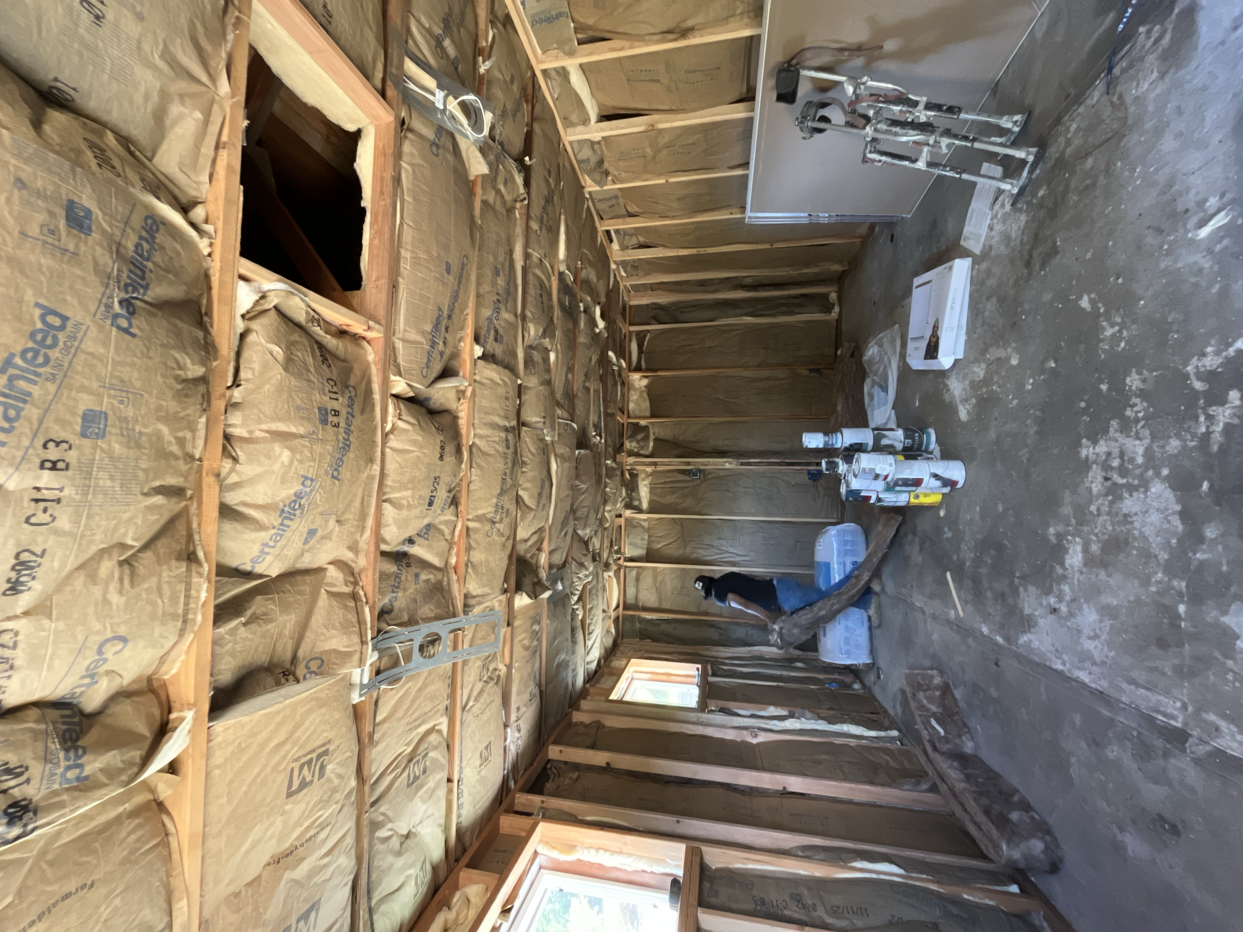 A construction site with wooden framing, insulation, and unfinished walls. There are various cans and tools on the floor, and a person working in the background.