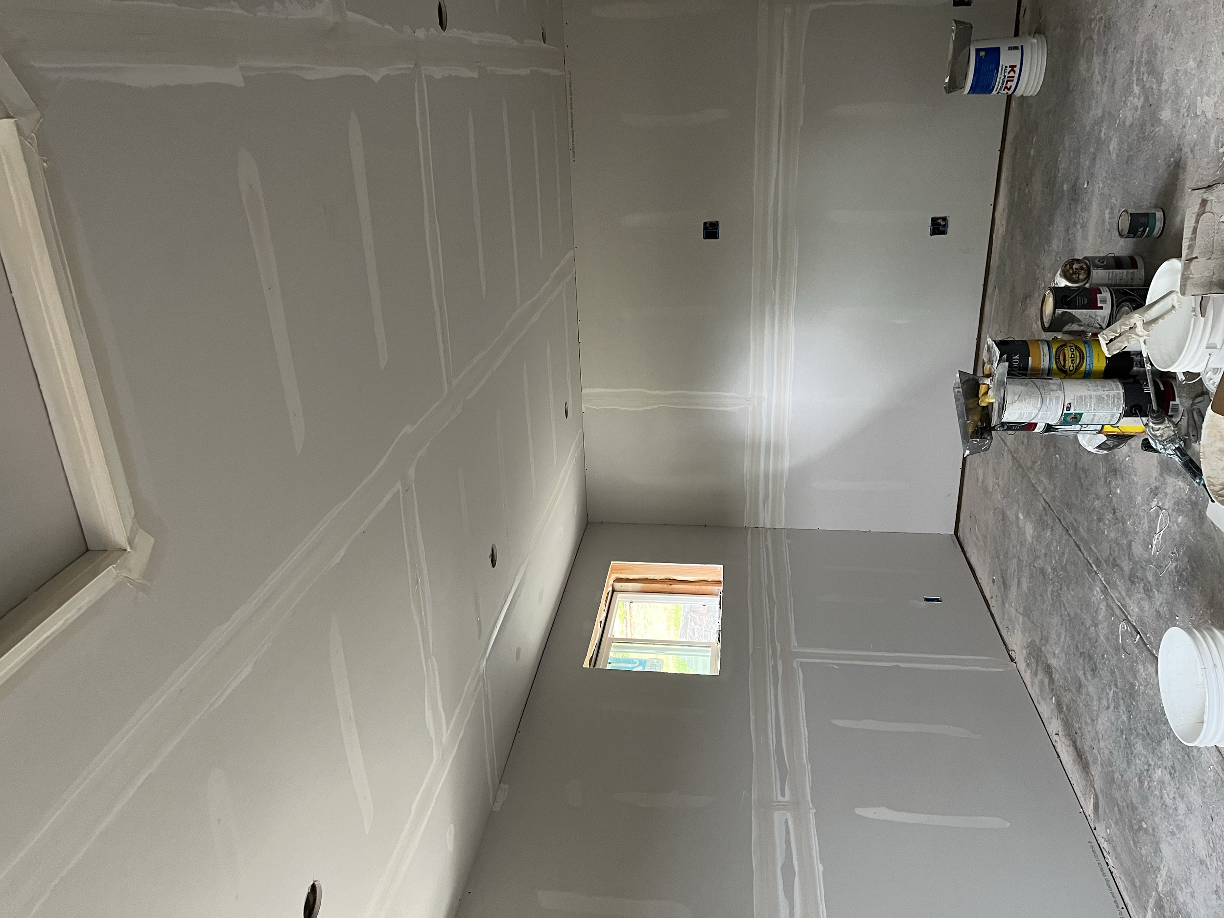 A room under renovation with drywall installed on the walls and a few electrical outlets without cover plates. The floor is unfinished, showing concrete, and there are various cans of paint or primer and construction supplies scattered on the floor. 
