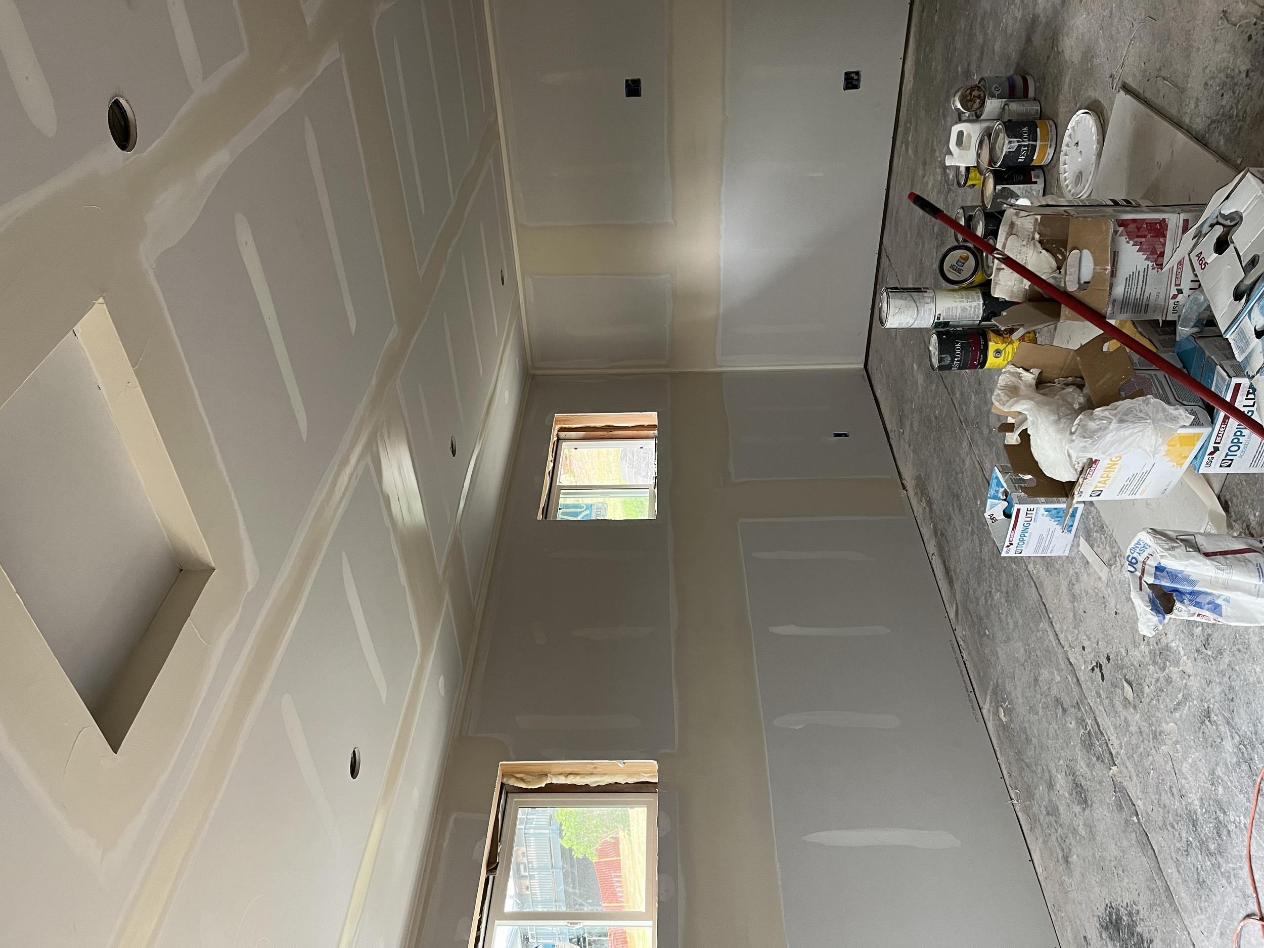 Interior of a room under construction with drywall installed on the walls and ceiling, with electrical outlets and light fixtures not yet put in. Construction supplies, paint cans, and tools are on the floor.