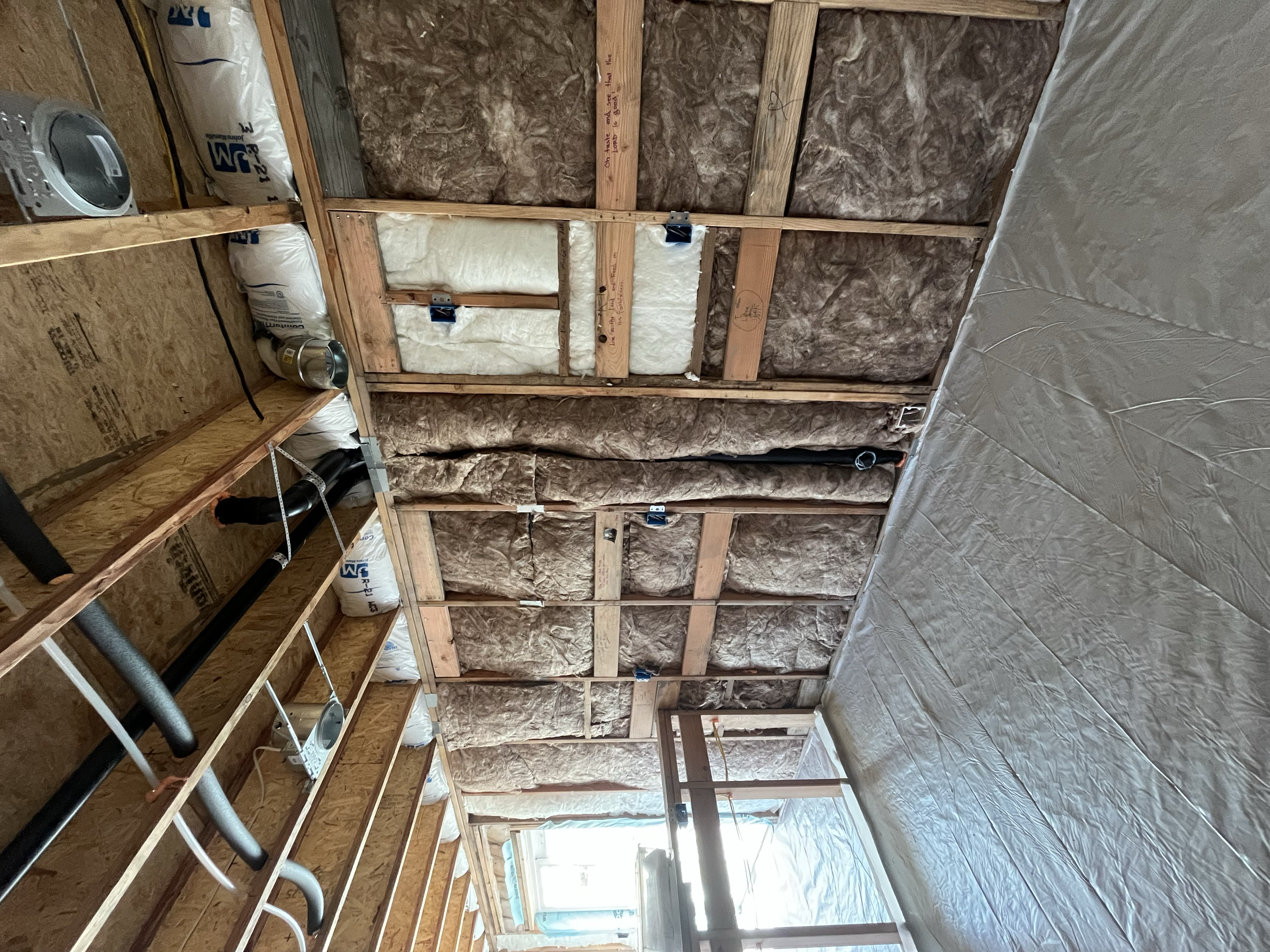 The inside of a building under construction or renovation showing wooden framing, insulation, and partially finished walls, with some tools and electrical wiring visible.