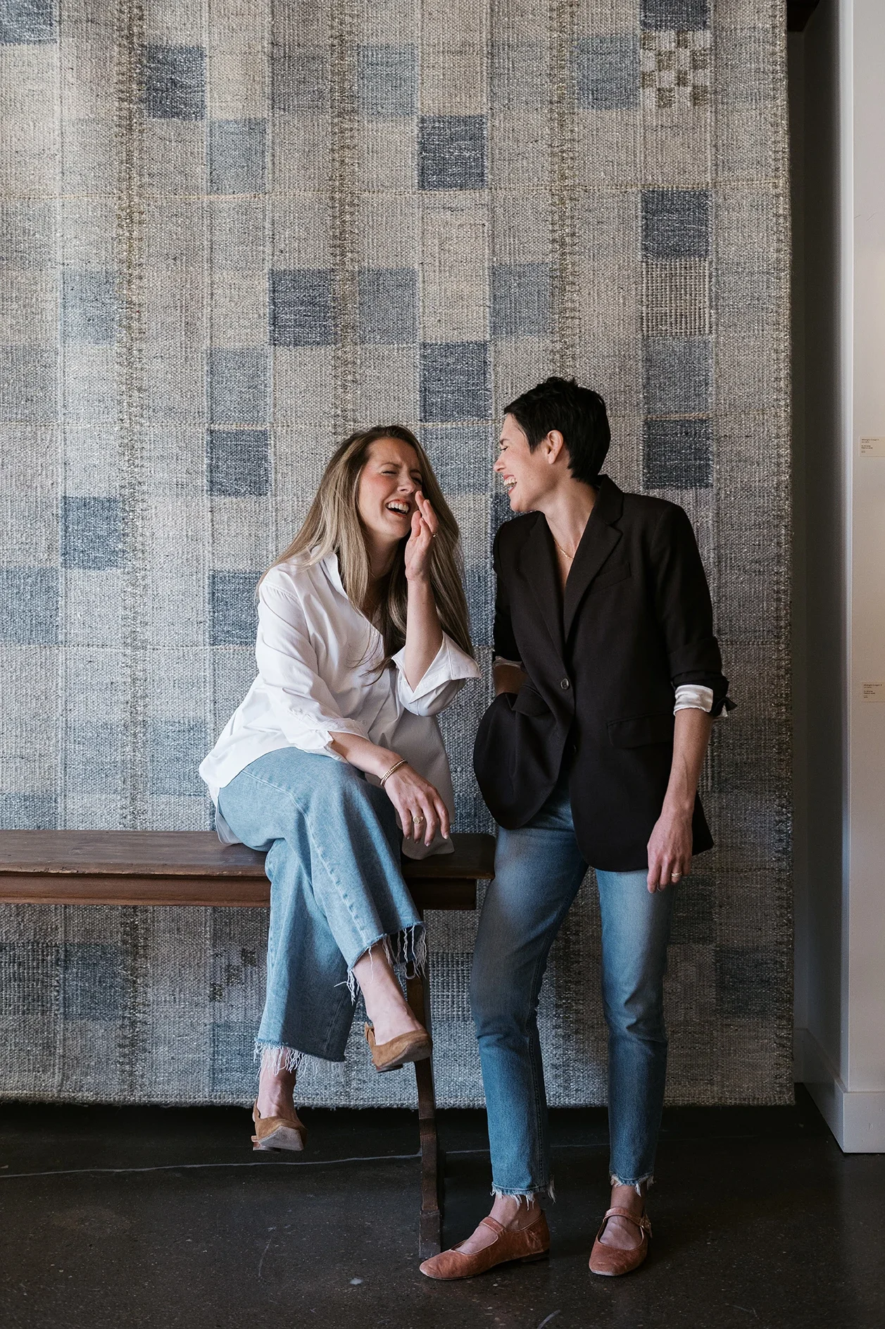 Two women are laughing and talking in an art gallery. One woman is sitting on a bench, wearing a white shirt and jeans, and the other is standing, wearing a black blazer and jeans. They are positioned in front of a textured, patterned wall.