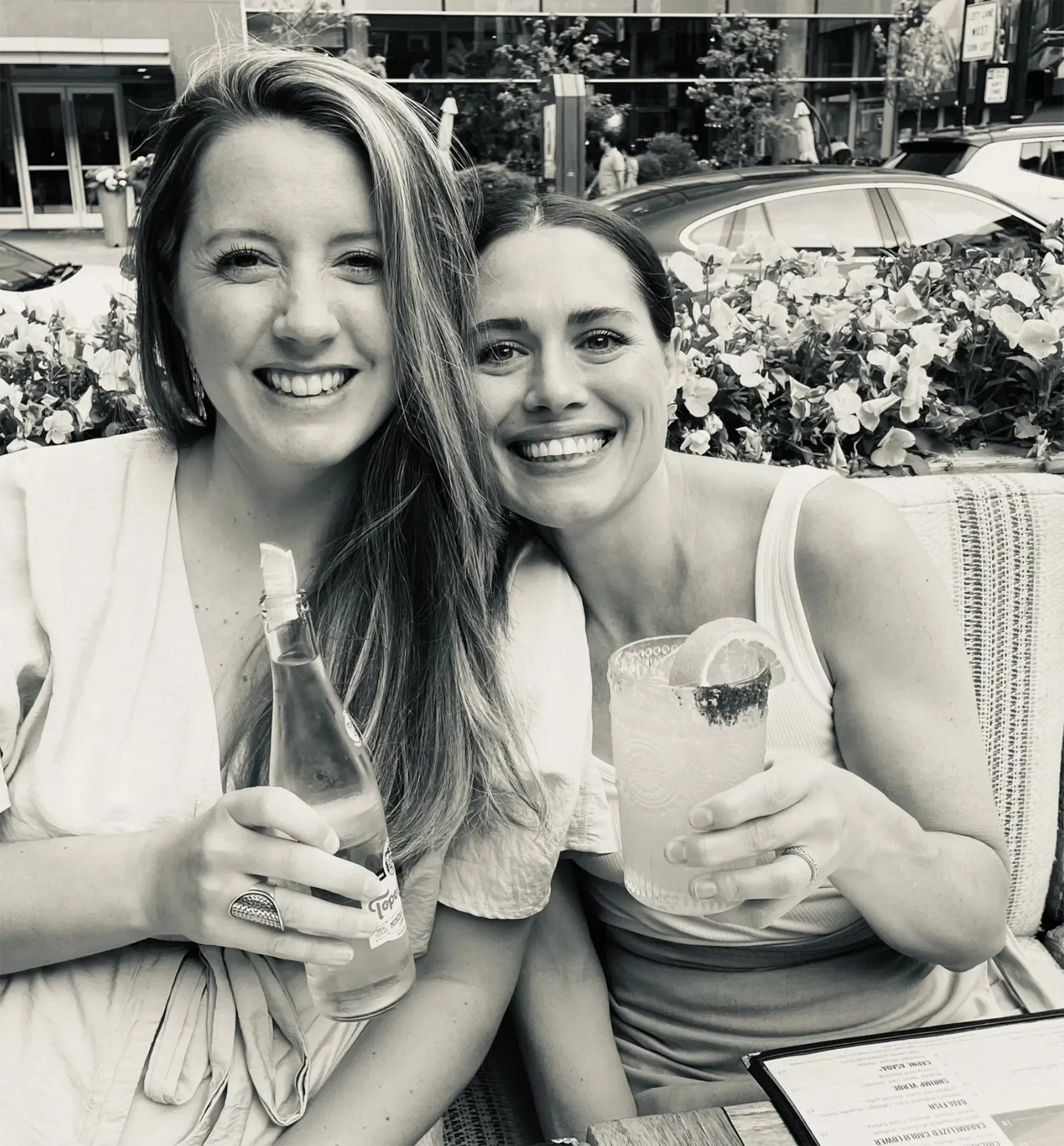 Two smiling women sitting at an outdoor table with drinks and a menu, in an urban setting with parked cars and pedestrians in the background.