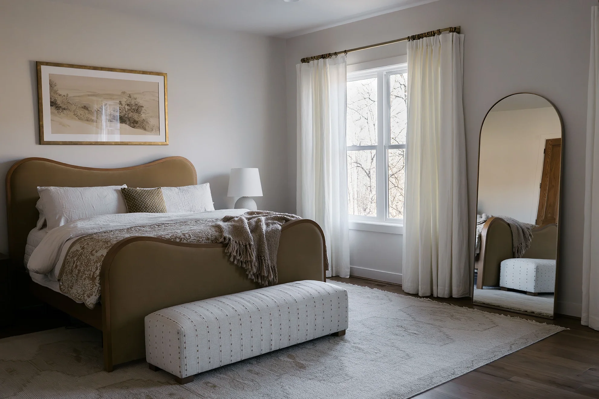 A cozy bedroom with two beds, a tufted ottoman at the foot of one bed, a white lamp on a side table, a large window with white curtains, and a floor mirror reflecting part of the room. There is a framed landscape picture above one bed.