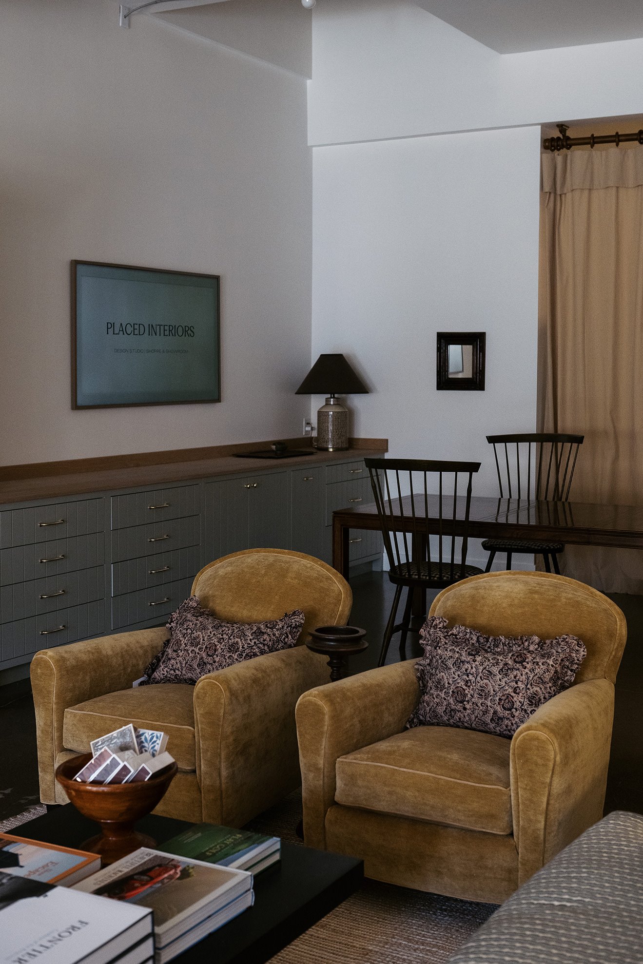 Interior of a living room with beige armchairs, decorative pillows, a wooden table, a gray cabinet, wall art, and curtains.
