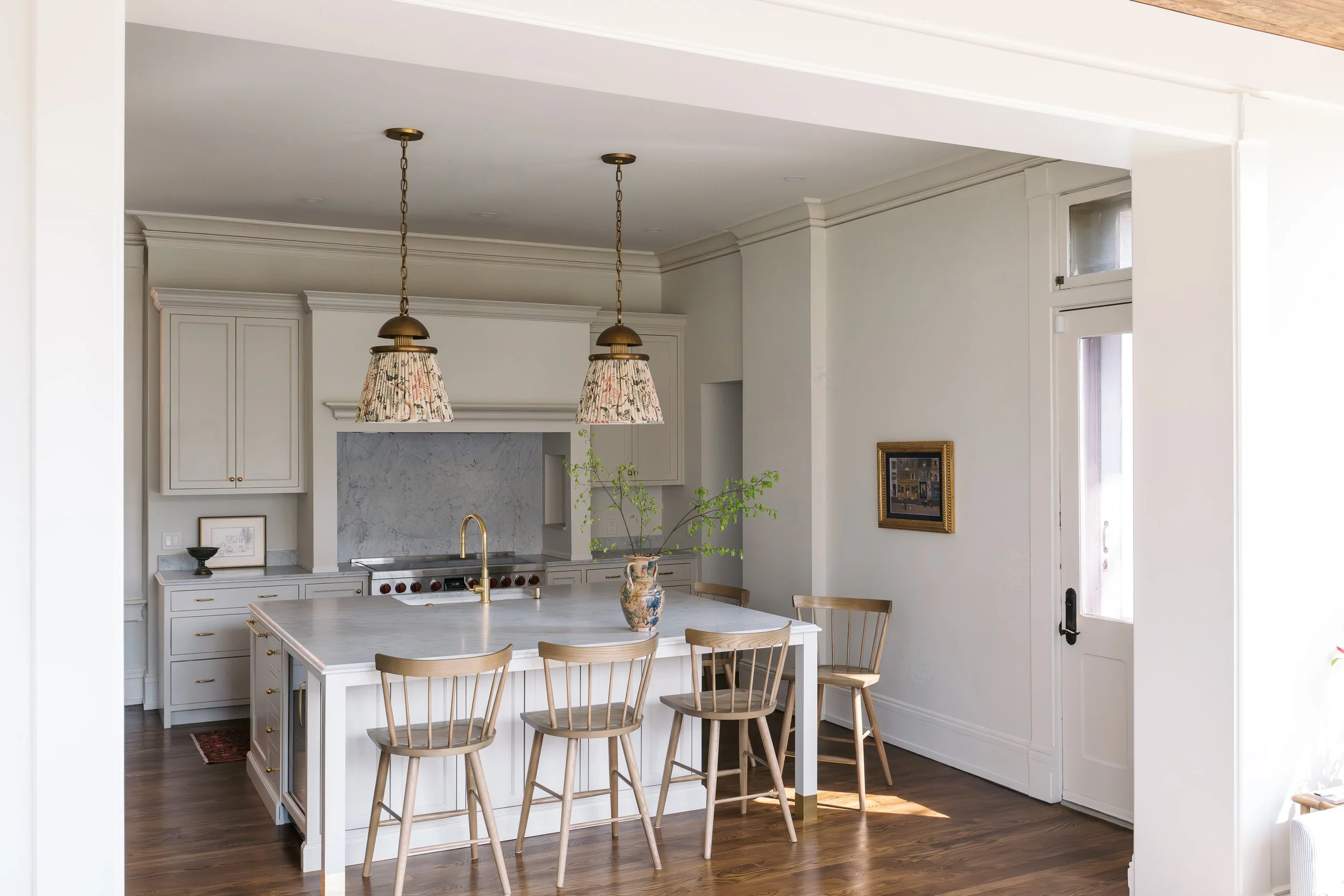 A modern kitchen with white cabinetry, a marble backsplash, a white island with a gold faucet, and hanging floral pendant lights. Wooden chairs surround the island, and a small window brings in natural light.