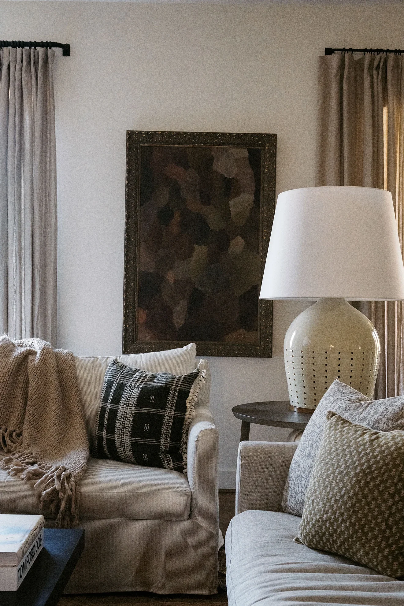 Living room with a cream-colored sofa, a black and white plaid throw pillow, a beige throw blanket, a side table with a white lamp, abstract framed art on the wall, and beige curtains.