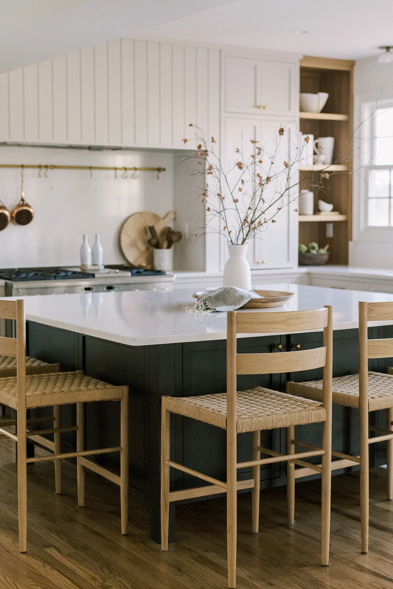 Modern kitchen with black island and natural wood chairs, white cabinetry, open shelves with white dishes, and a window in the background.