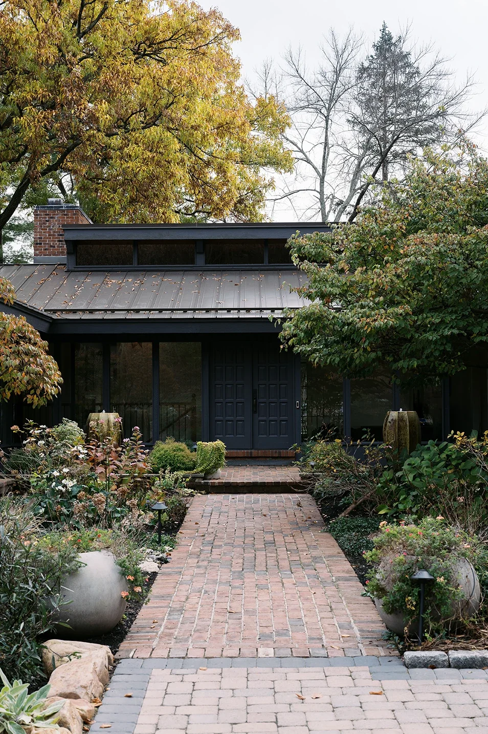 A brick pathway leads to the entrance of a modern house with black framing and large windows, surrounded by lush landscaped garden with trees and potted plants.