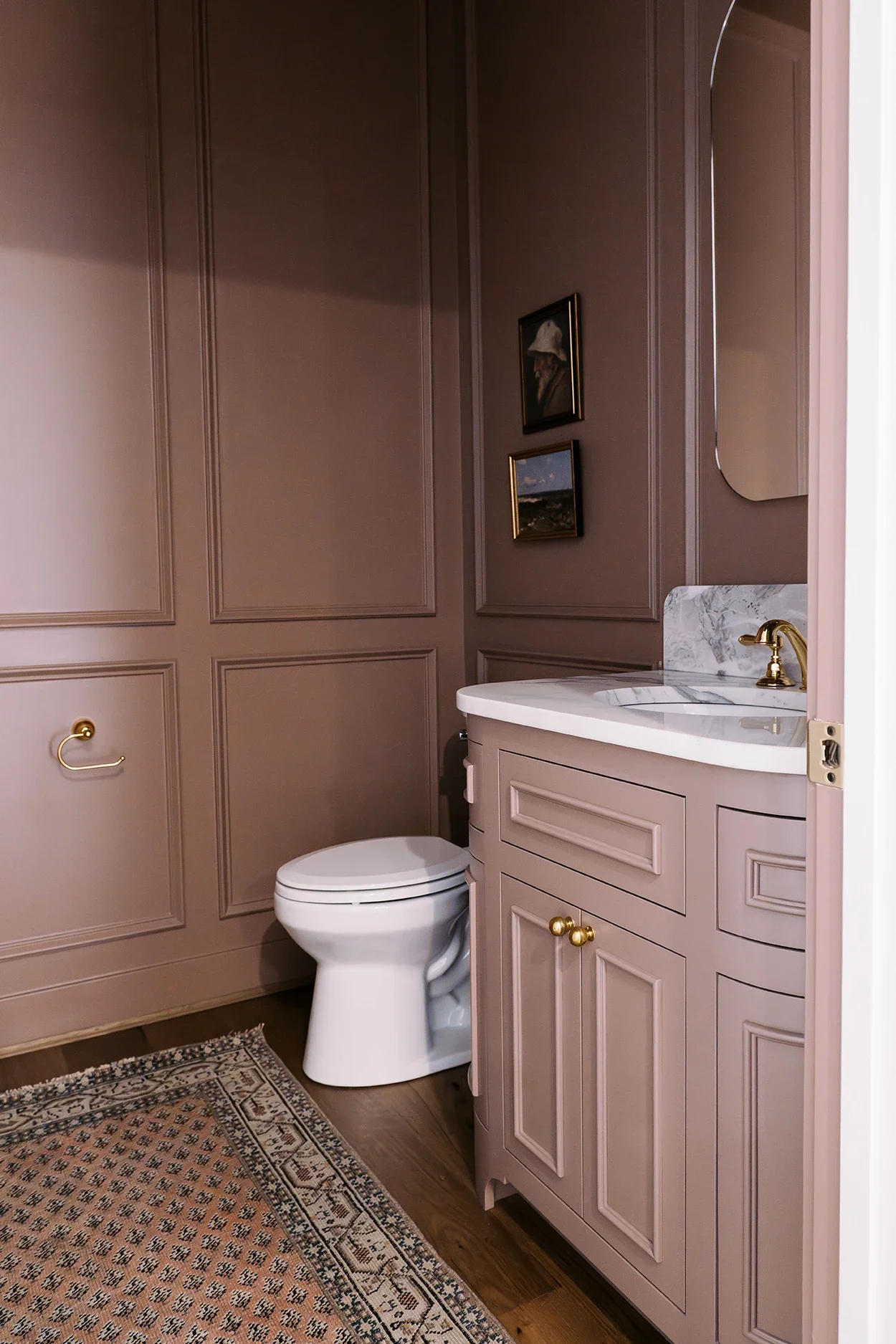 A bathroom with taupe-paneled walls, a white toilet, a beige vanity with a marble countertop, and a gold faucet. Two framed paintings hang on the wall, and there is a mirror and patterned rug on a wooden floor.