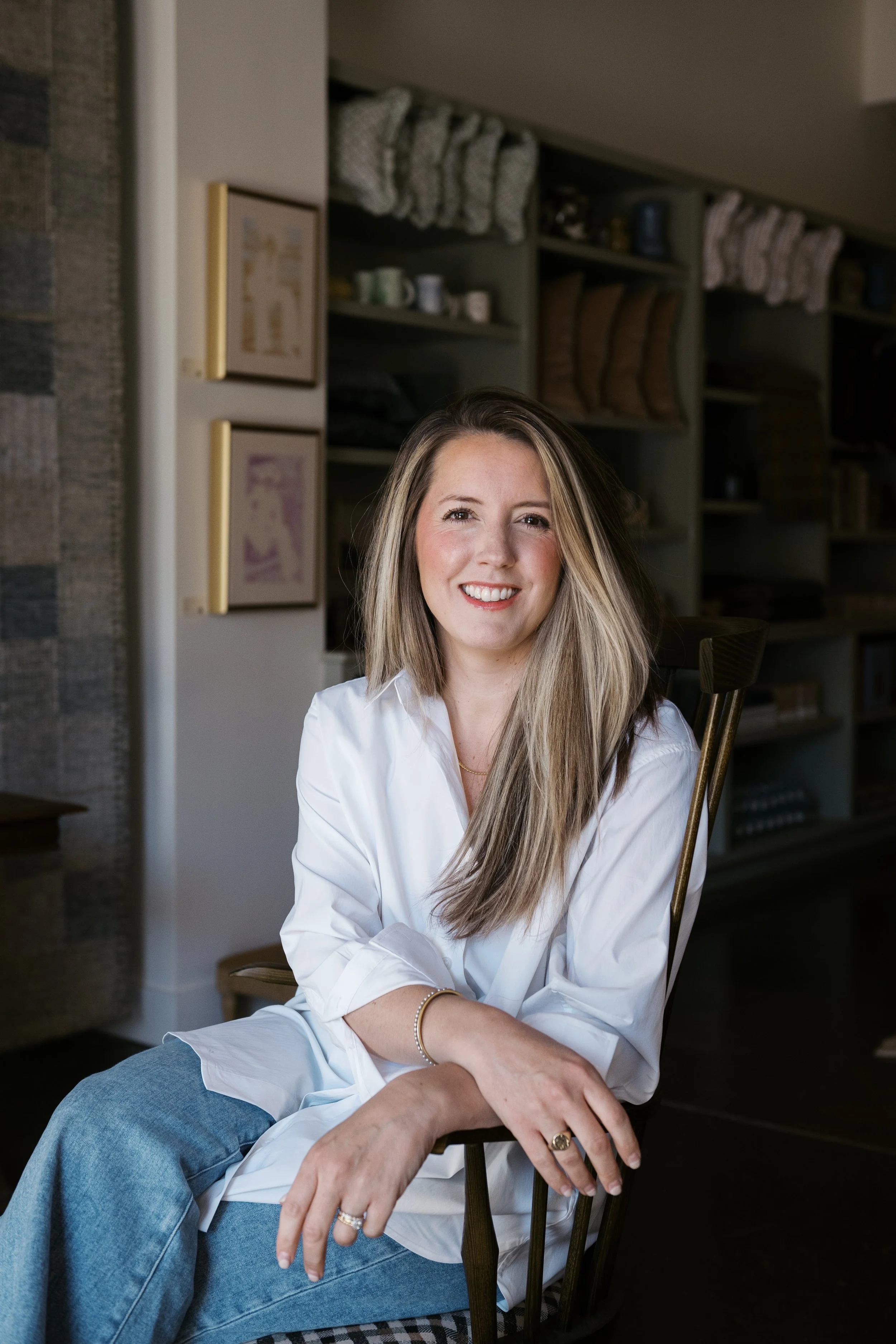 A woman with long blonde hair smiling while sitting on a wooden chair in a cozy kitchen or dining room. She is wearing a white shirt and blue jeans, with multiple rings and a gold bracelet. Behind her are open shelves with cups, bowls, and towels, and a brick wall is partially visible on the left.