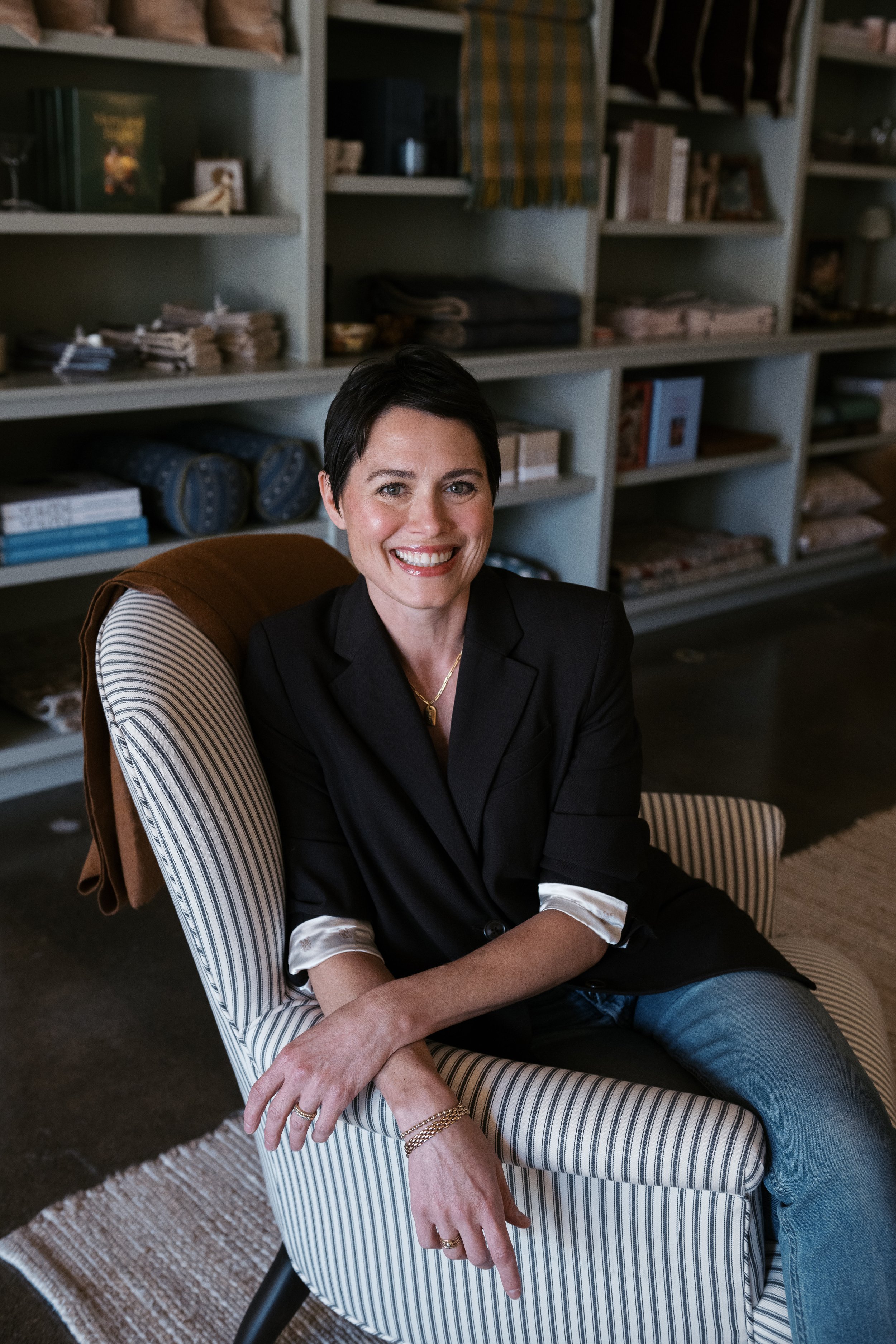 A woman with short dark hair and a bright smile, wearing a black blazer and light-colored shirt with rolled-up sleeves, sits on a striped armchair in a room with bookshelves filled with books and folded fabrics.