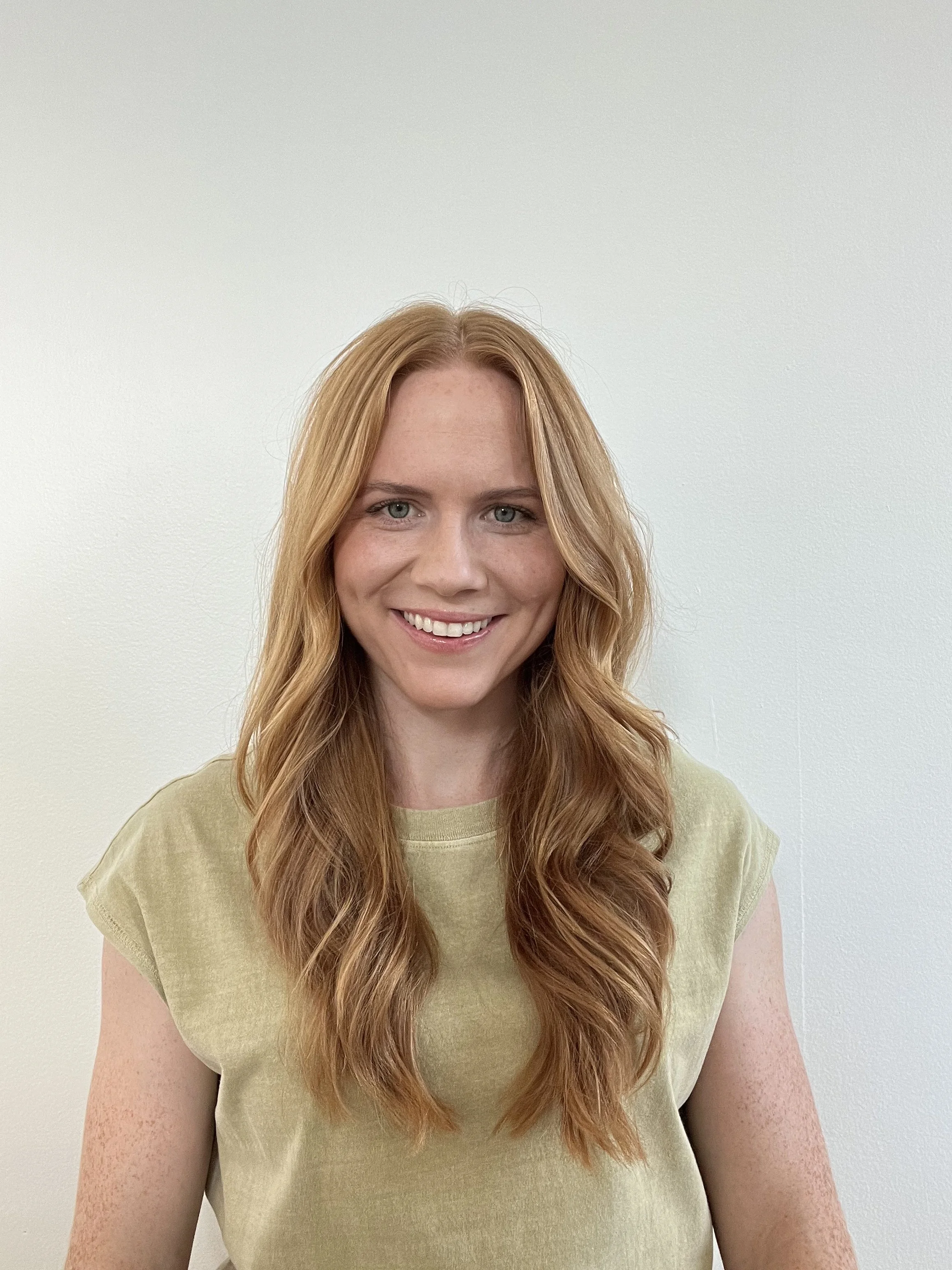 A young woman with long, wavy red hair, light skin with freckles, smiling and wearing a light yellow T-shirt, standing against a plain white wall.
