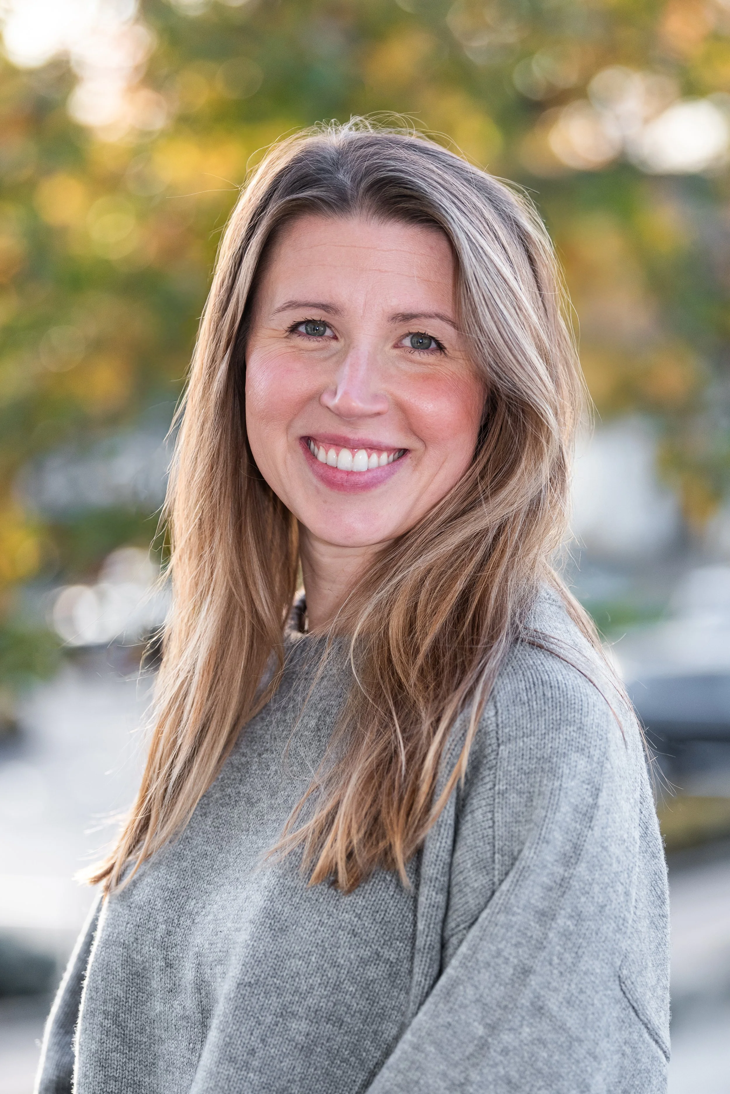 Close-up of a smiling woman with long blonde hair, wearing a gray sweater, outdoors with a blurred background of autumn foliage.