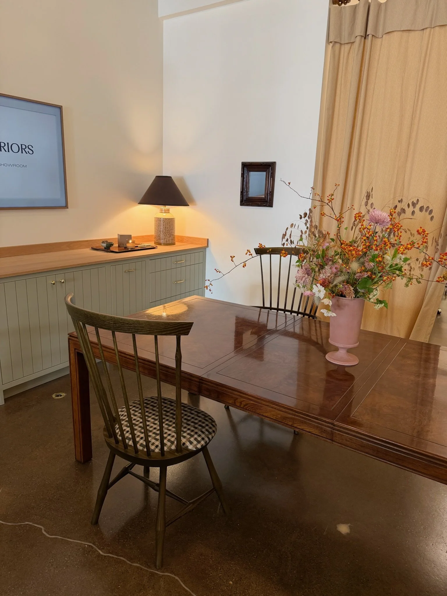 A room with a wooden dining table and a black chair with a checkered cushion. A large pink vase with flowers is on the table. There's a beige cabinet with a lamp and decorative items on top. A wall with curtains, a small mirror,  are visible.