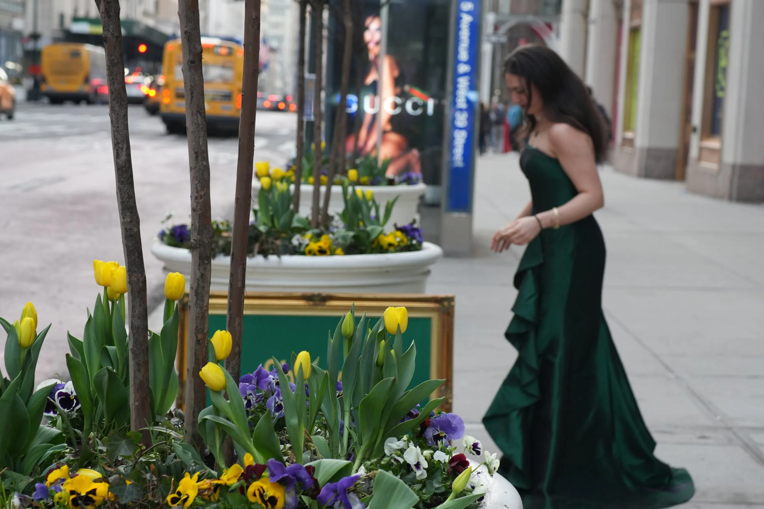 City street scene with a woman in a green dress looking at flowers in a planter, yellow tulips and purple and white flowers, with blurred vehicles and buildings in the background.