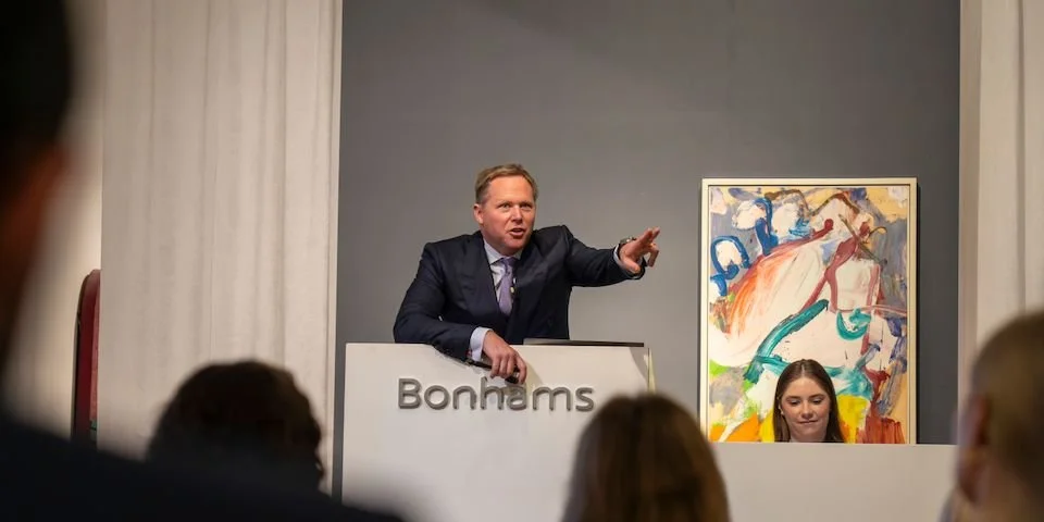 Auctioneer at Bonhams auction house speaking to an audience, with a colorful abstract painting behind him.