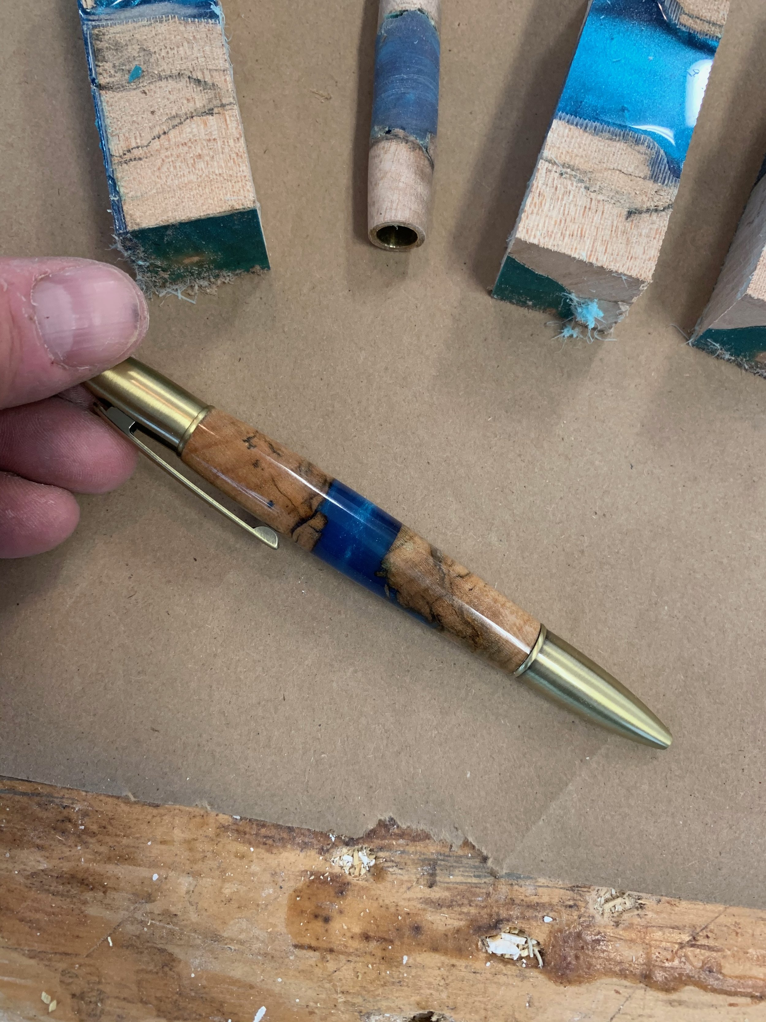 A wooden surface, an open pen, a woodworking drill bit, and two rectangular pieces of wood placed on a craft board. The pen has a blue and lighter wood grain pattern and the woodworking drill bit is cylindrical with a rough, worn appearance.