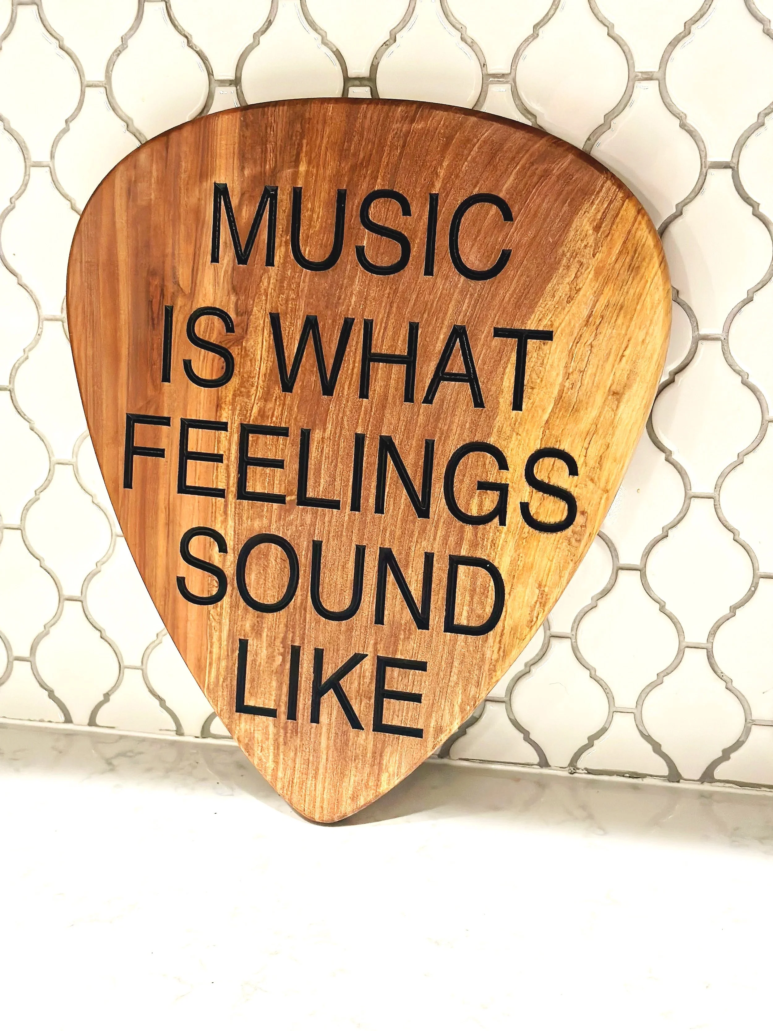 A wooden sign shaped like a guitar pick with the words "Music is what feelings sound like" written in black text, leaning against a white tile wall with a patterned grout design.