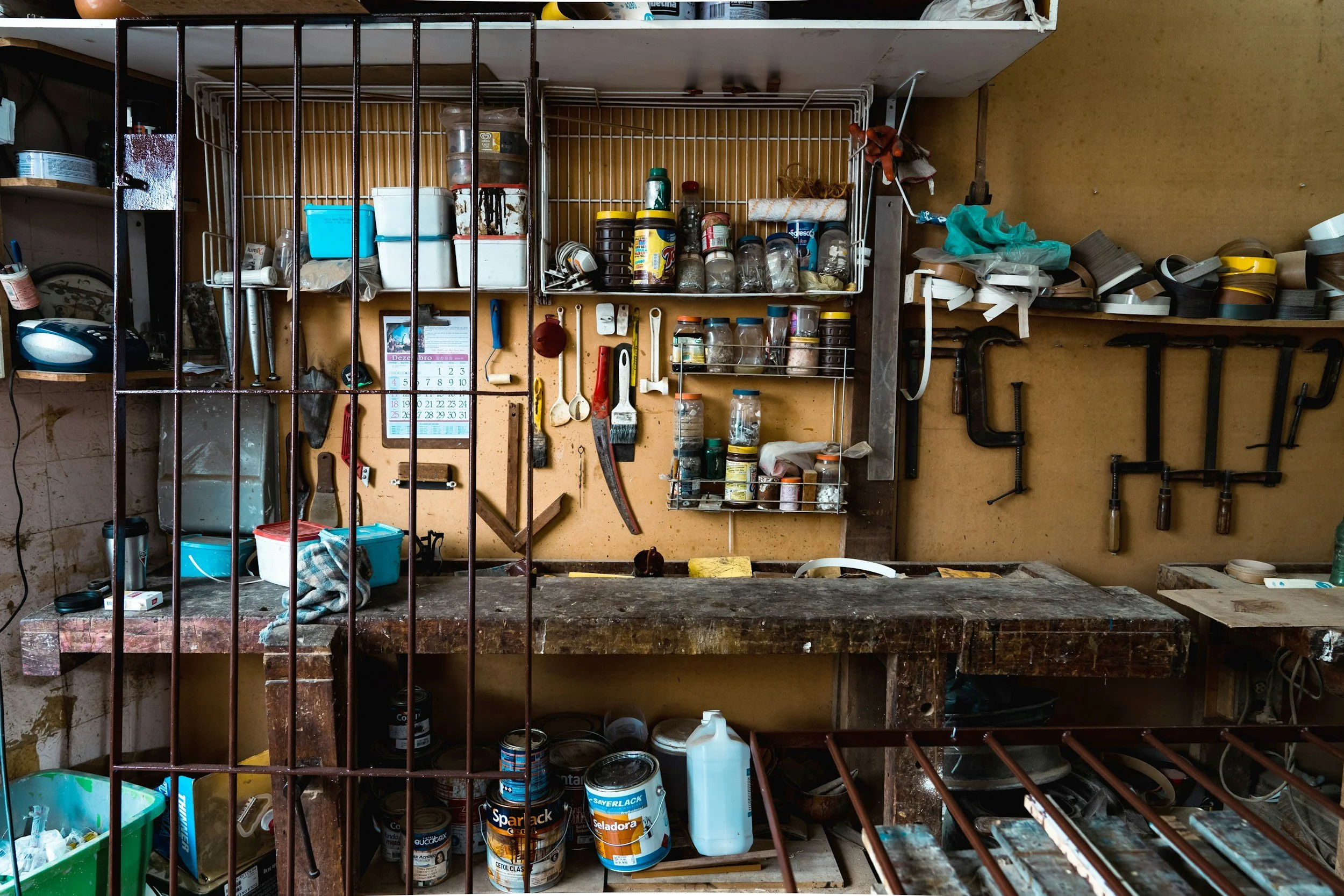 View of a cluttered workshop or storage room with shelves holding paint cans, jars, tools, and various supplies, partially obscured by metal bars in the foreground.