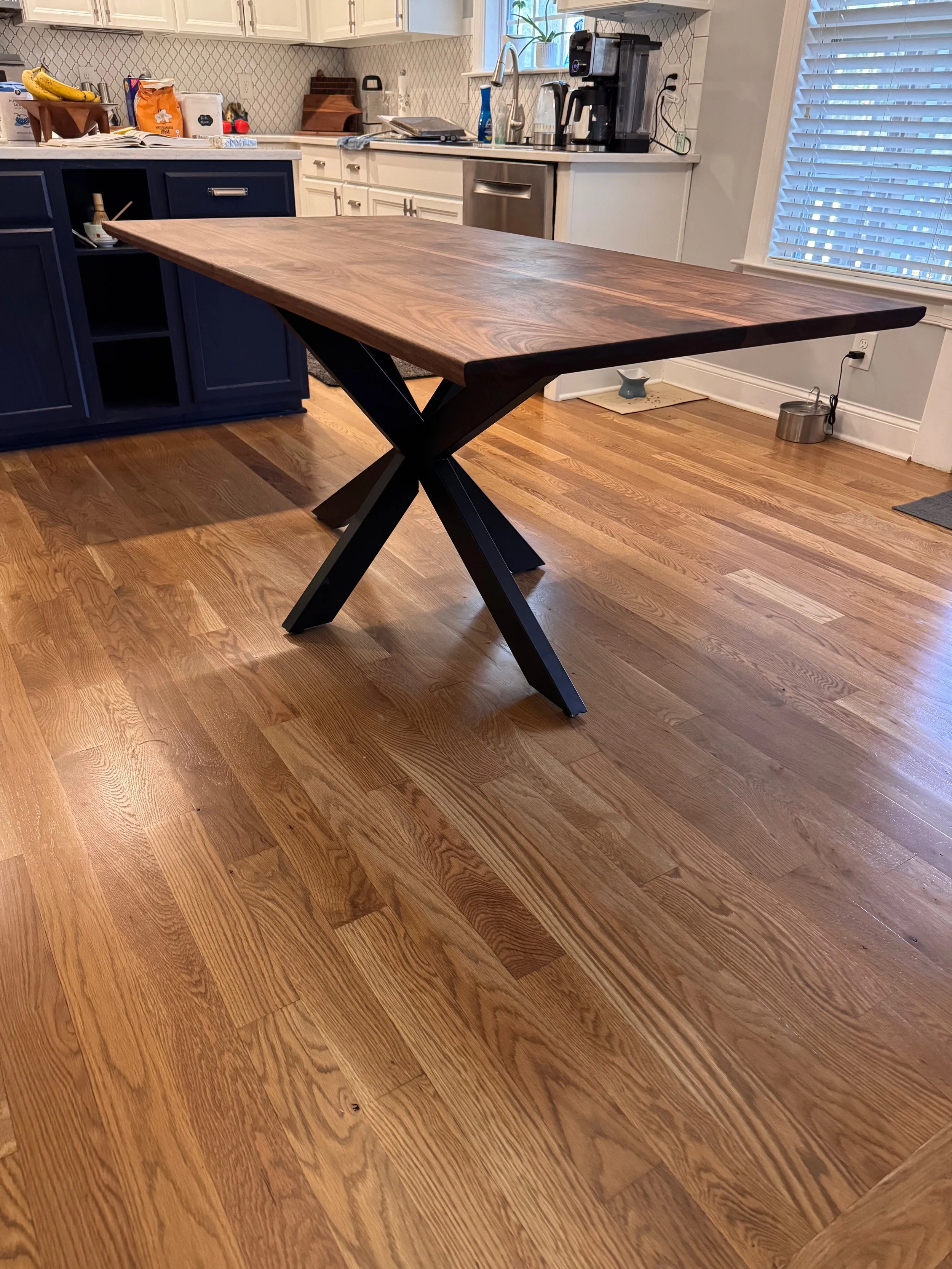 A wooden dining table with black X-shaped legs in a kitchen with hardwood floors, white cabinets, and various kitchen appliances and items on the counter.