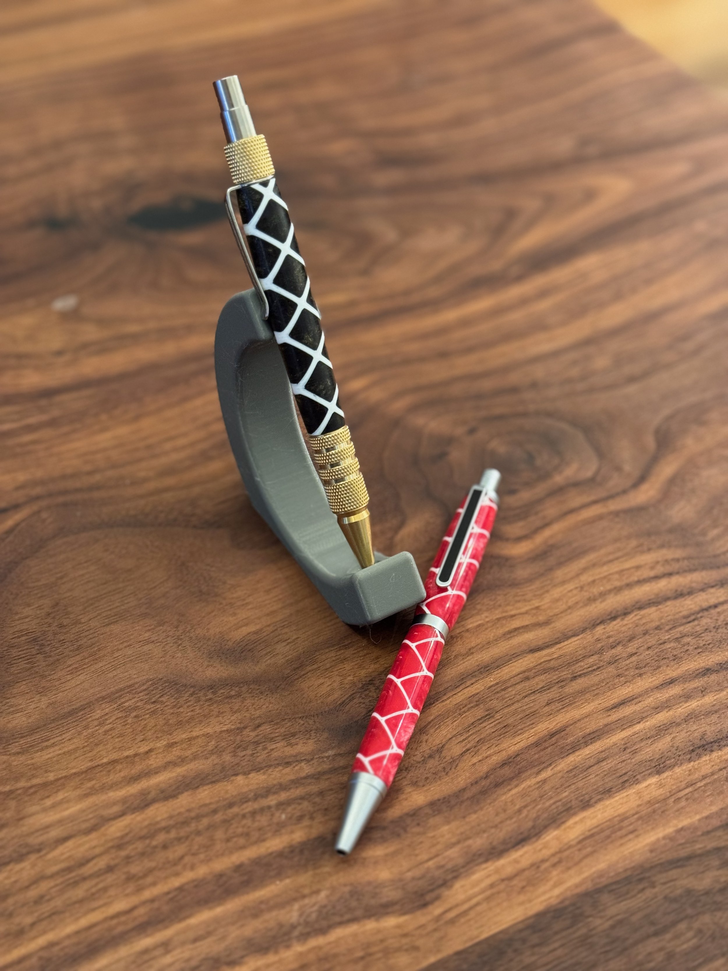 Close-up of a tactical pen with a black and white pattern, mounted on a gray stand on a wooden surface, with additional pens in the background.